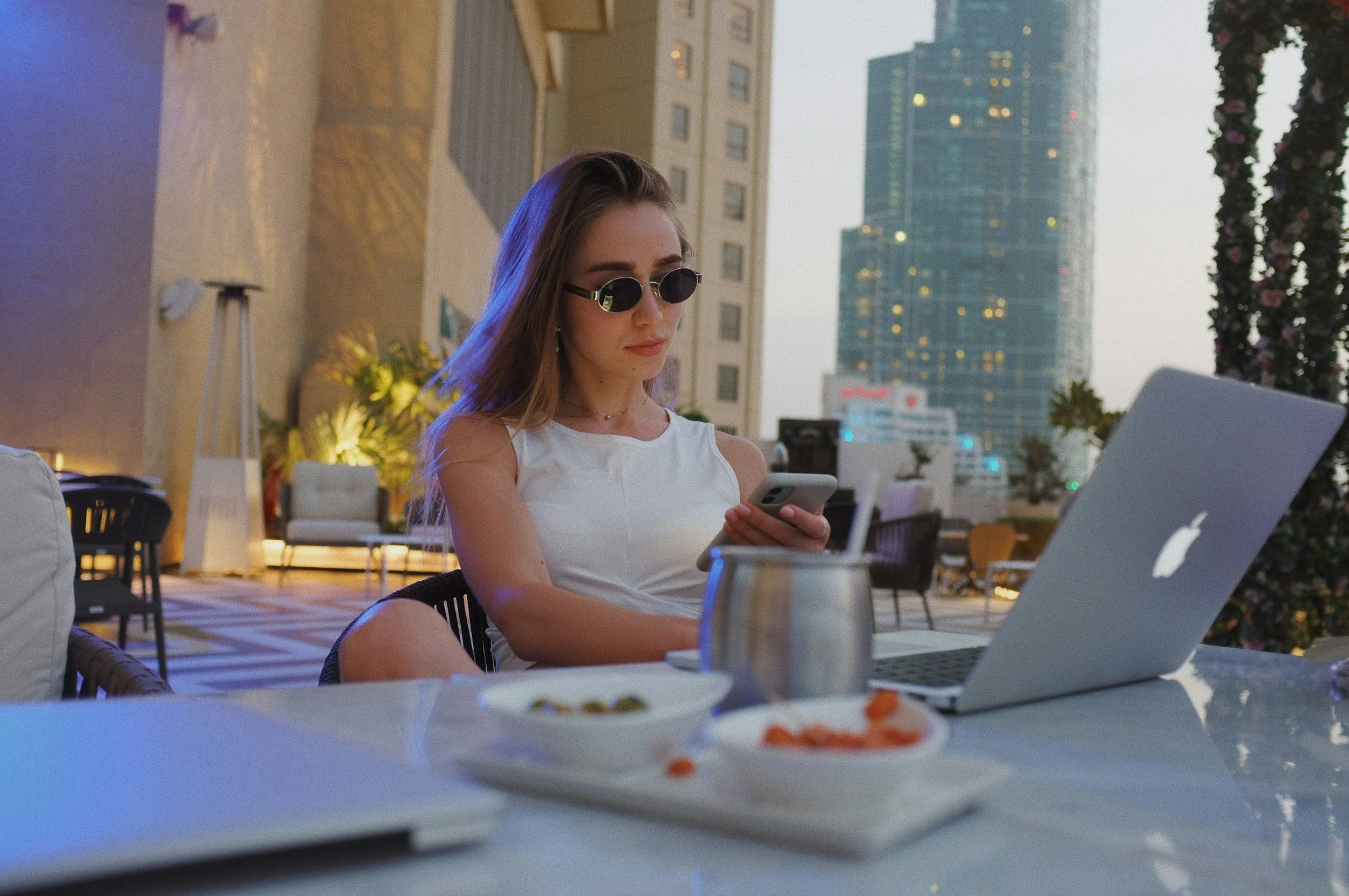 Young woman wearing sunglasses and a sleeveless white top sitting at an outdoor terrace, looking at her phone, with a MacBook, plates of food, and a metal cup on the table, with city buildings in the background during evening.