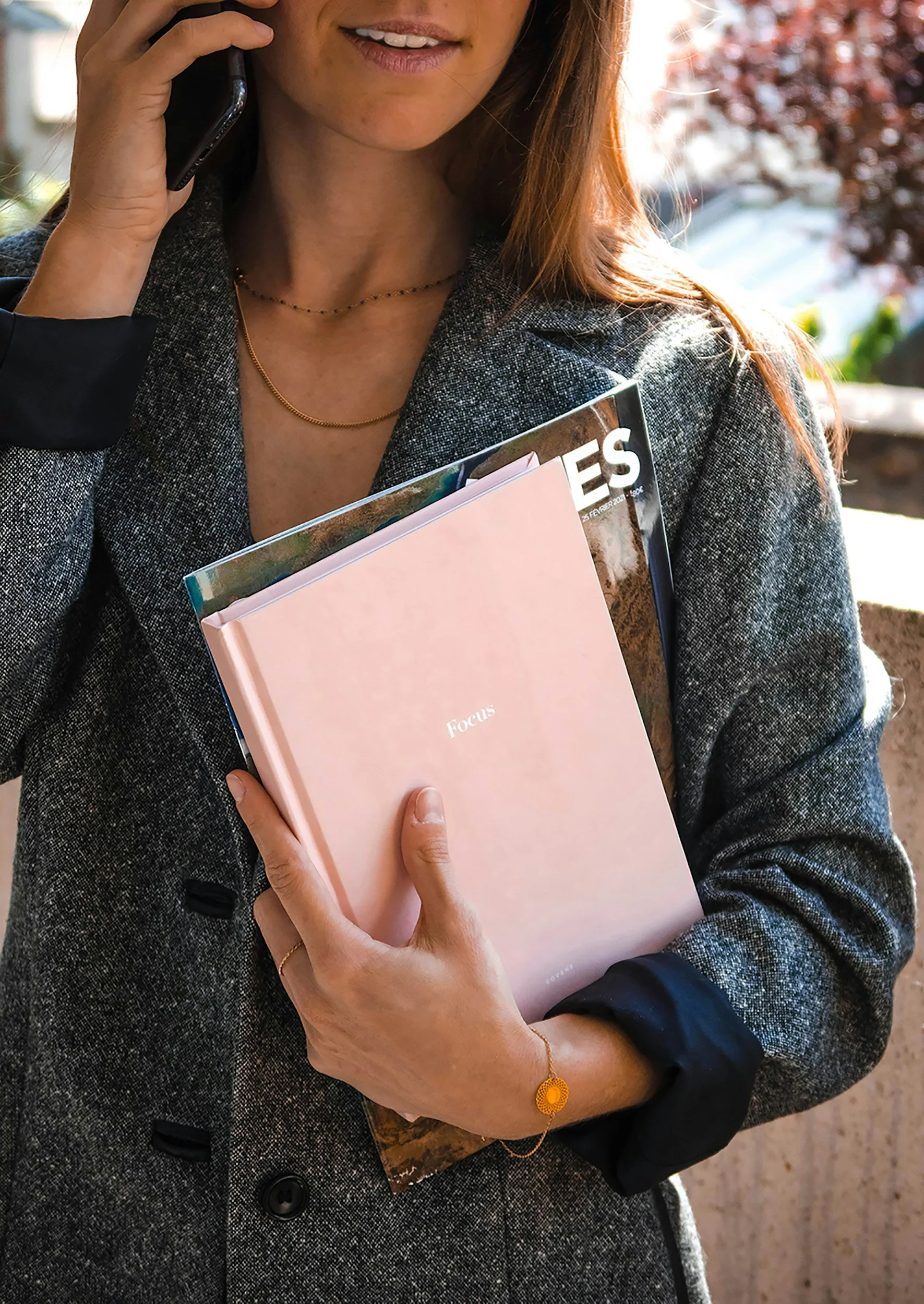 A woman holding a smartphone to her ear, wearing a gray blazer, gold jewelry, and holding a pink book labeled 'Focus.' She is outdoors with sunlight and blurred trees in the background.