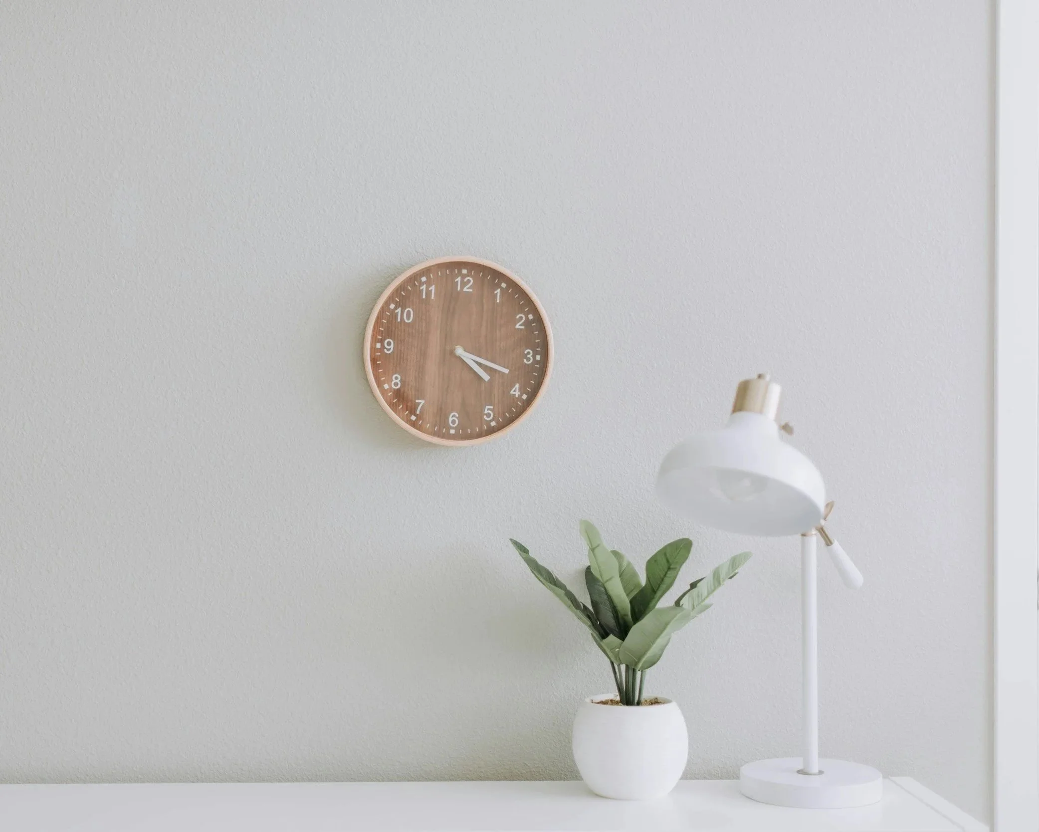 White desk with a potted plant, a white desk lamp, and a wood clock on a plain white wall.