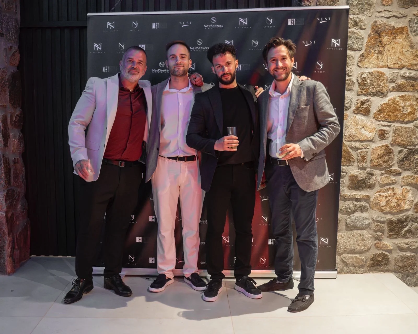 Five men dressed in business and casual attire standing together, holding drinks, at an event with a black backdrop and stone wall background.