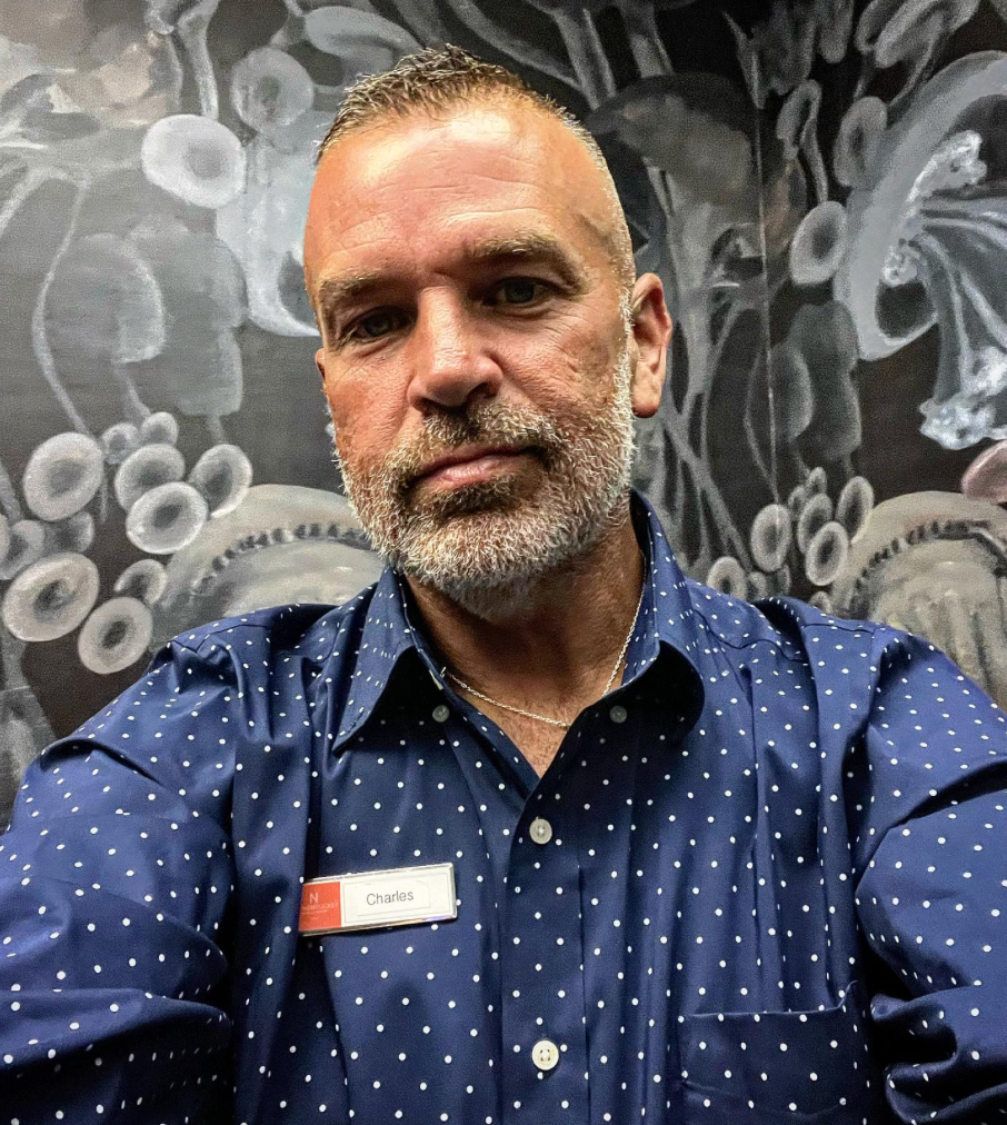 A man with a beard and short hair taking a selfie in front of a gray mural with abstract designs. He is wearing a navy blue shirt with white polka dots and a name tag that reads "Charles."