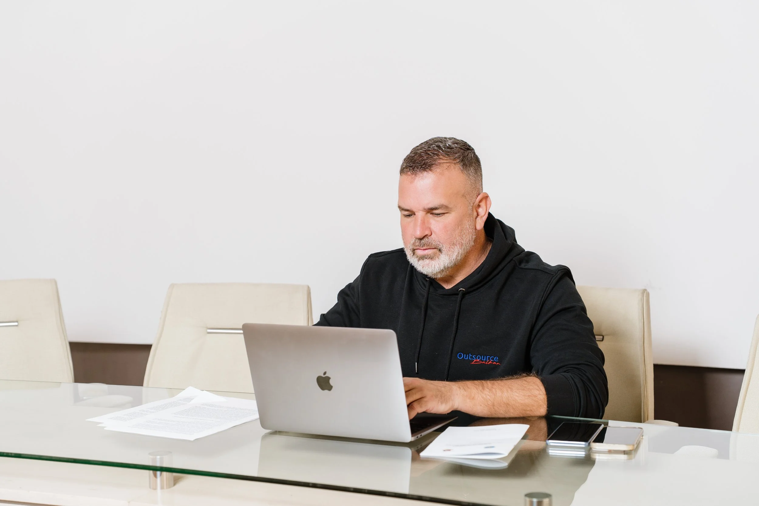A man with short hair and a beard working on a laptop at a white table in a room with white walls, surrounded by papers and two smartphones.
