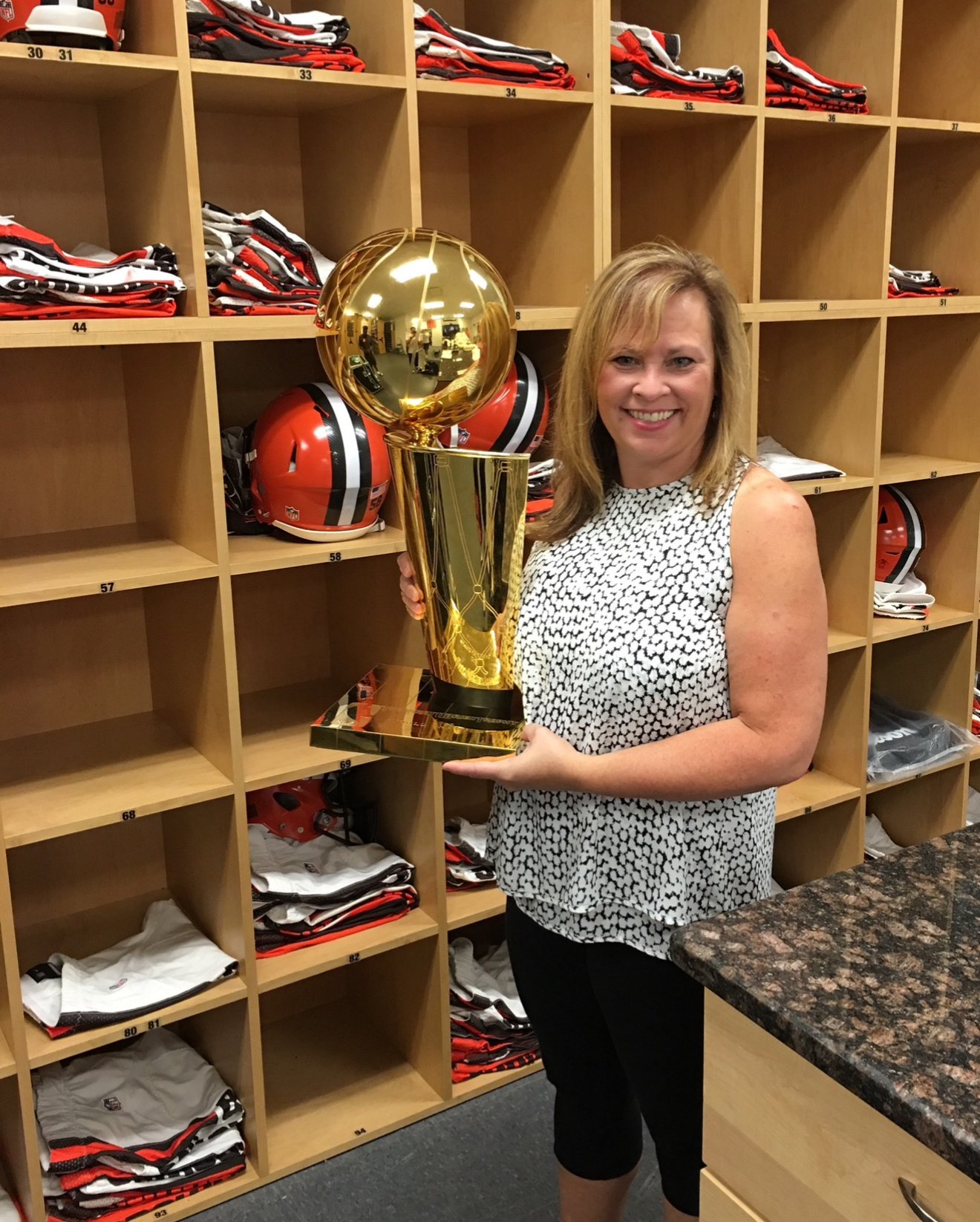 A woman smiling and holding an NBA trophy in front of a wooden locker with NFL jerseys and helmets.