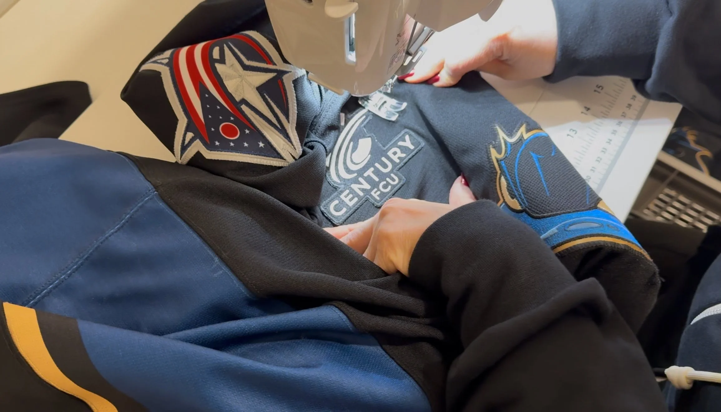 Close-up of a person sewing a sports jersey with the logos of the Columbus Blue Jackets and the Cleveland Monsters . The person is wearing a dark gray long-sleeve shirt, and a sewing machine is being used to stitch the fabric.