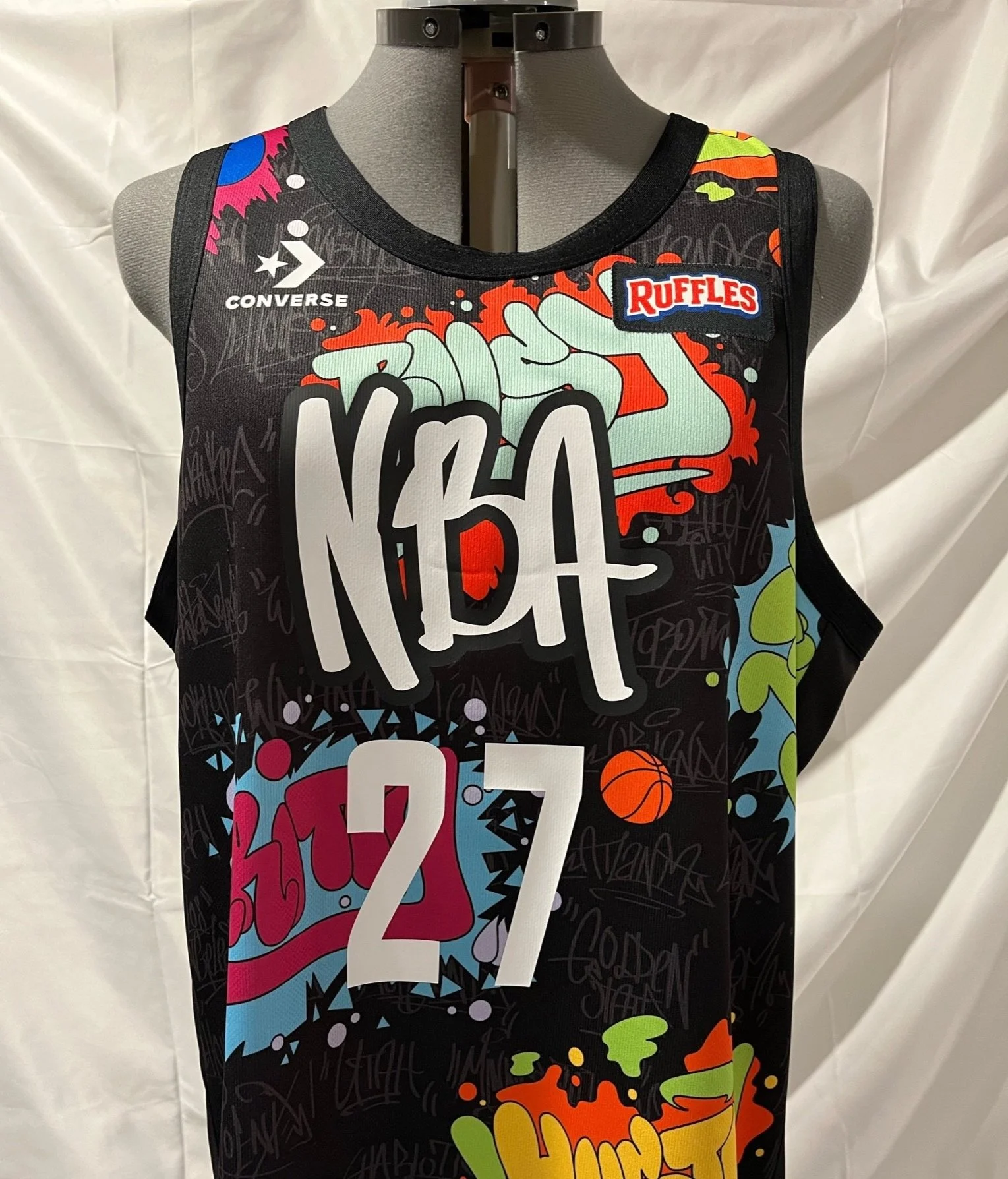 NBA All Star celebrity game jersey on a mannequin featuring graffiti-style artwork, large text, and various sponsor logos including Converse and Ruffles, with a black background and colorful swirling designs.
