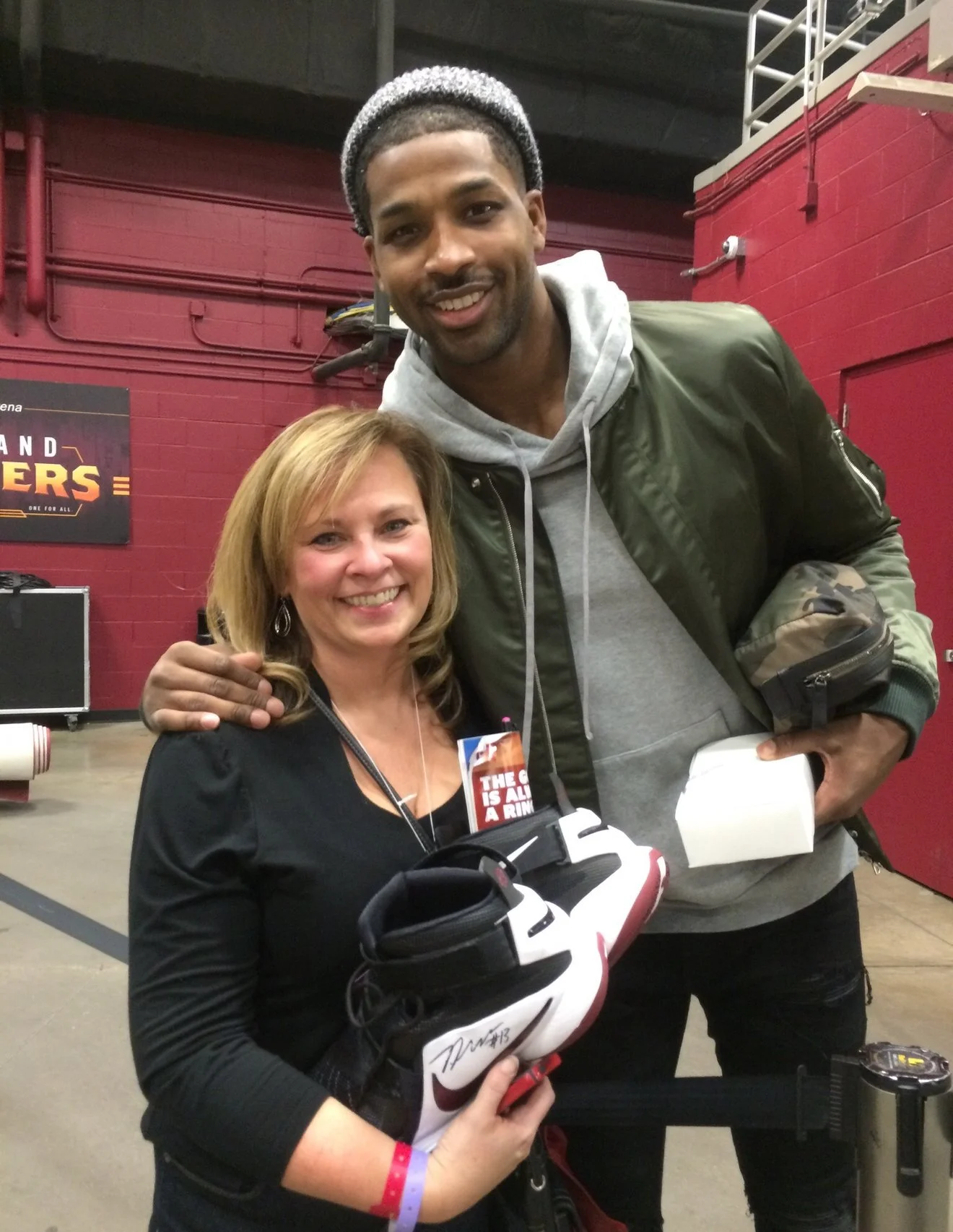 A woman and NBA player Tristan Thompson in a sports arena after a game. She is holding game worn shoes, with the background of red brick walls and a sign that reads 'HEROES'.