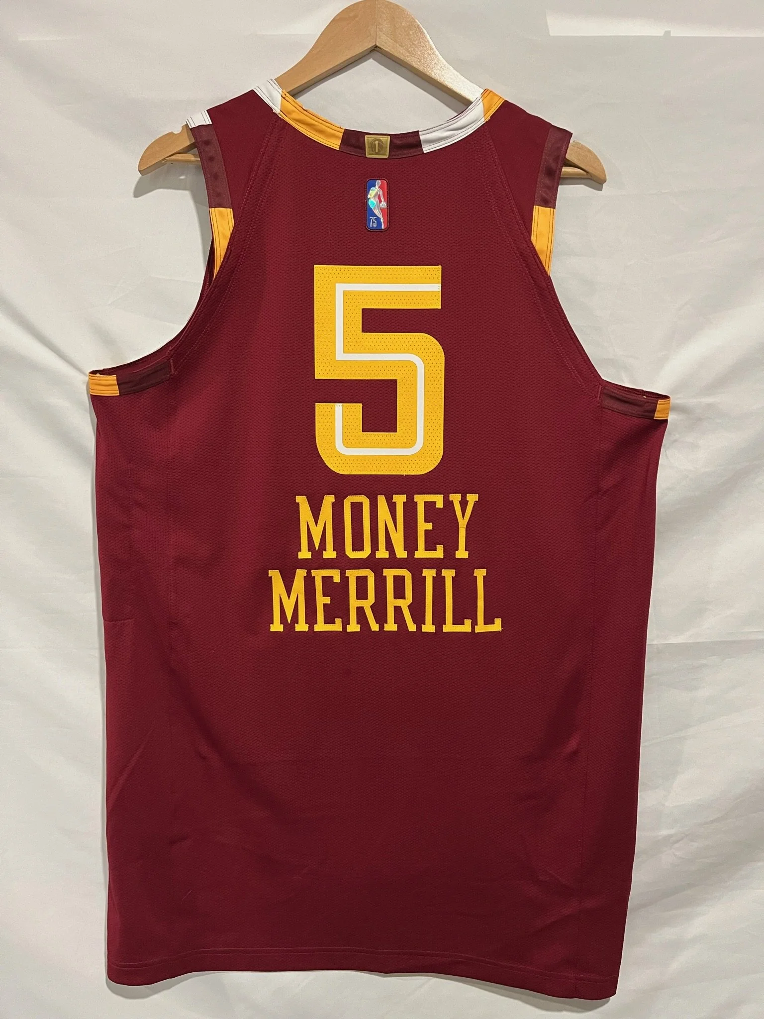 A maroon Cavs basketball jersey with gold and white accents hanging on a wooden hanger. The back displays the number 5 in gold and the name 'Money Merrill' in gold capital letters.