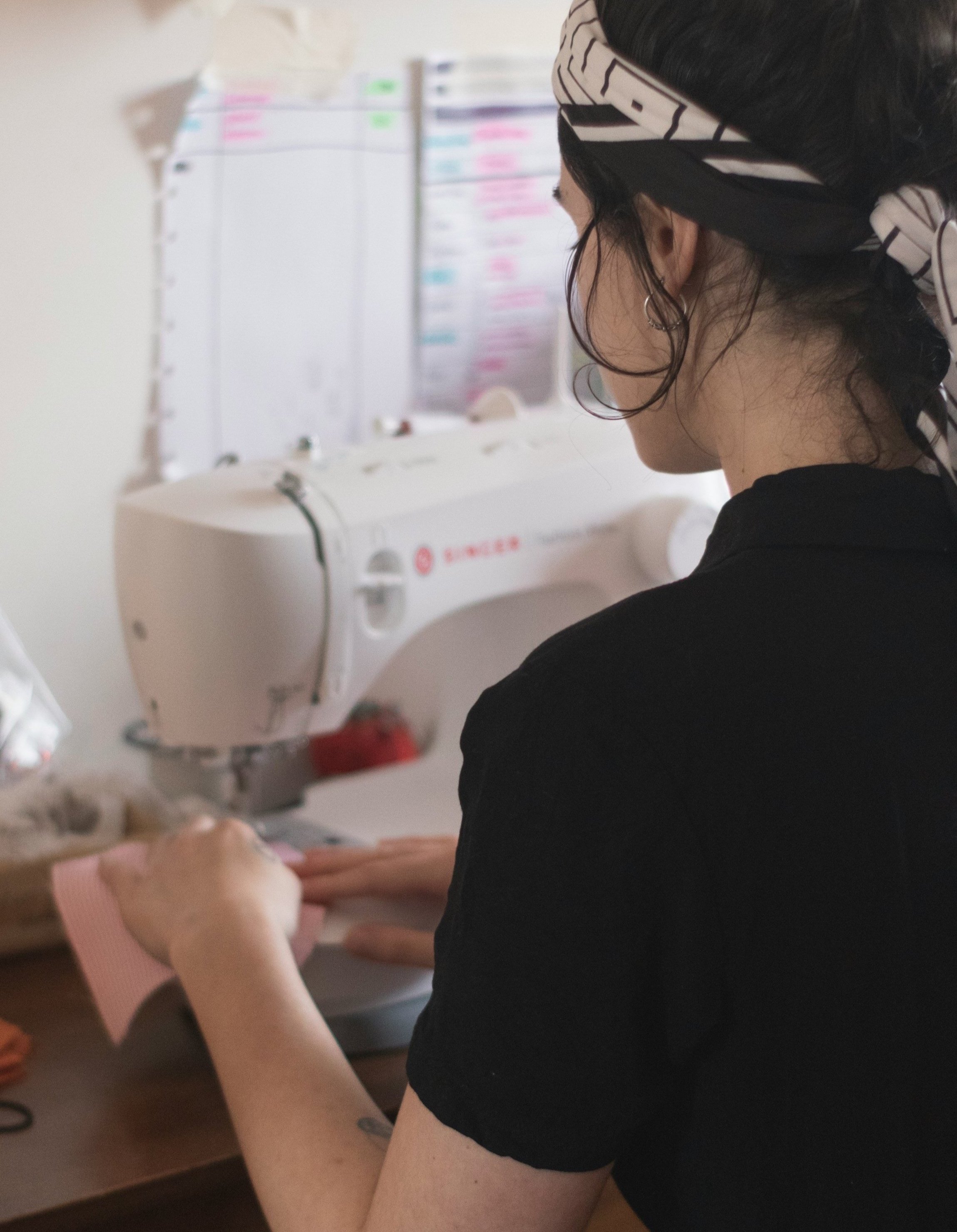 A woman with dark hair, wearing a striped headscarf, is working on a sewing machine. She is dressed in a black top and is focusing on her sewing project, which involves a piece of pink fabric. The background shows a wall with a whiteboard or a planning sheet with colored notes or markings.