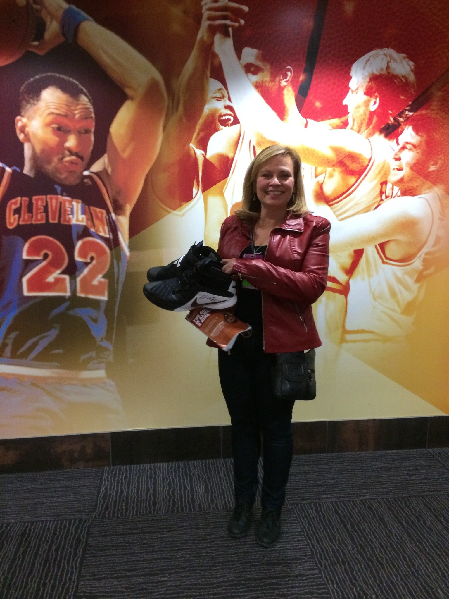 A woman standing in front of a large mural of a basketball game, holding a pair of shoes and smiling at the camera.