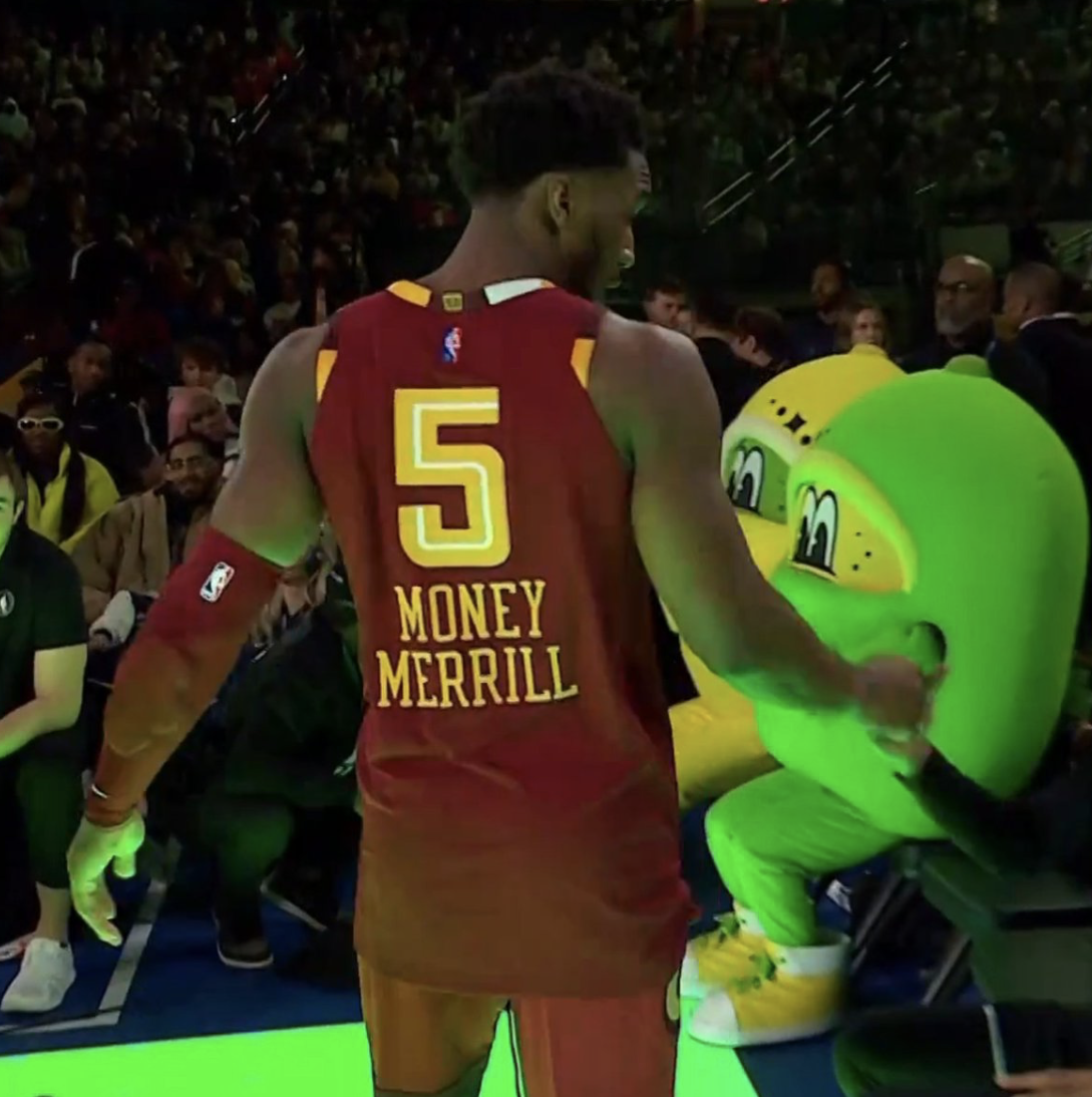 Donavon Mitchell wearing a jersey with the number 5 and the name Money Merrill is reaching toward a seat where a large green mascot is seated. The background shows an audience watching the game during a 3 point shooting competition.