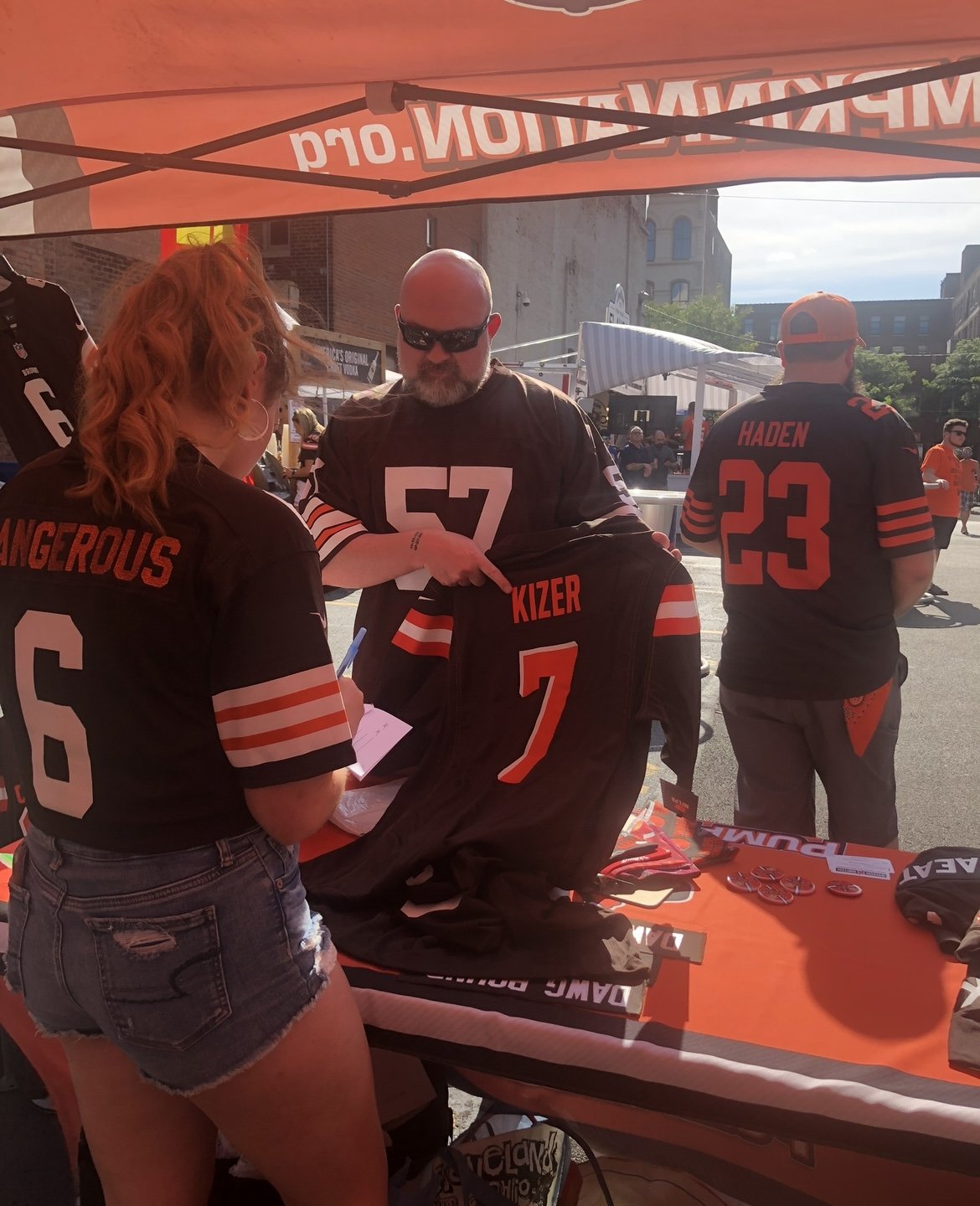 People at a sports merchandise booth, wearing brown football jerseys with orange and white accents, during an outdoor sports tailgate.
