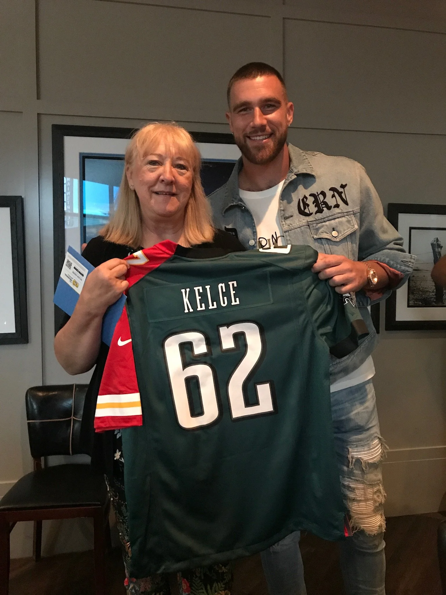 A woman with blonde hair holding a Kansas City Chiefs jersey with the number 62, standing next to a man with short hair and a beard, wearing a denim jacket and smiling.