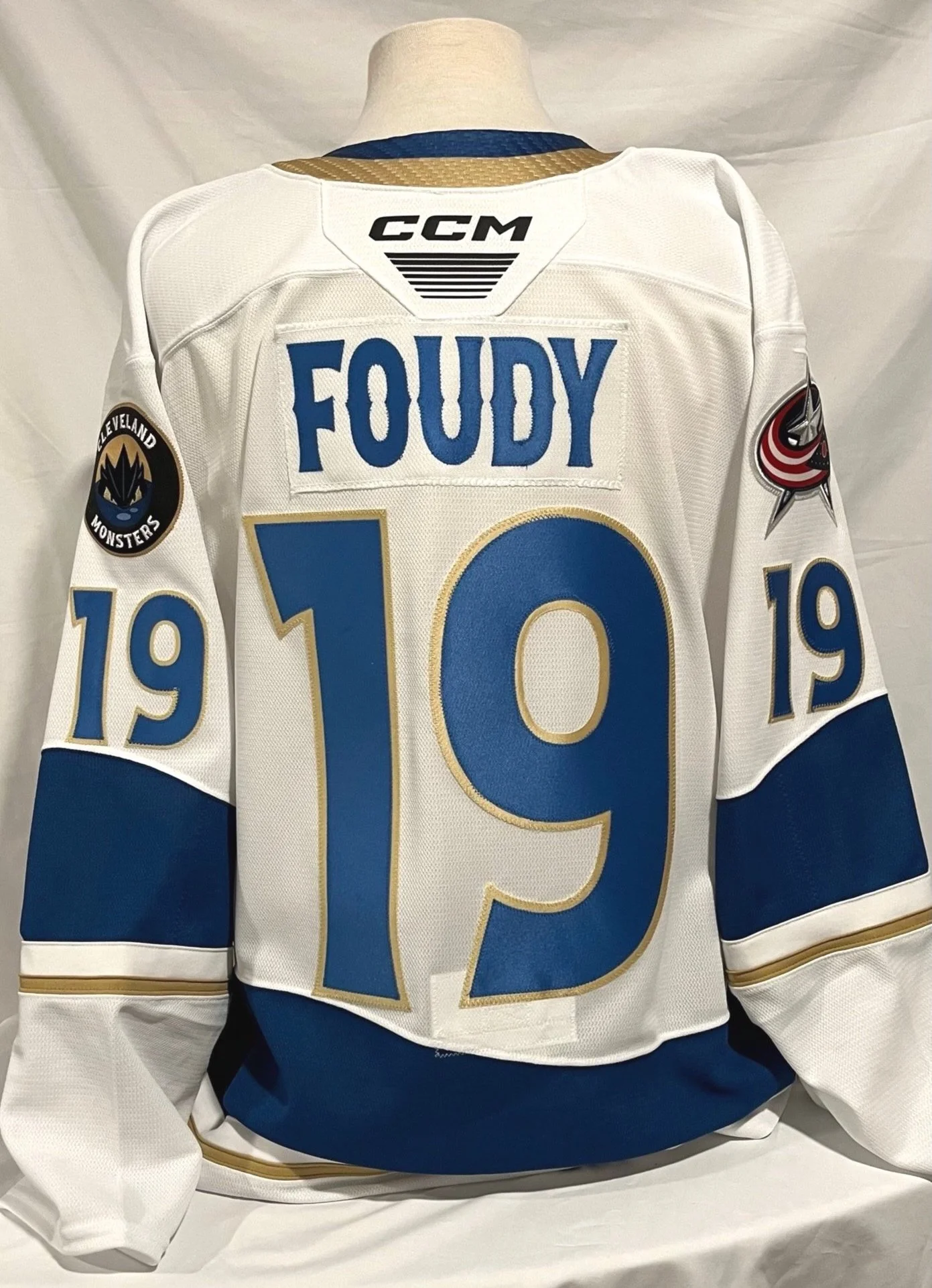 A white Cleveland Monsters hockey jersey with blue and gold accents. It features the name 'FOUDY' and the number '19' on the back, with a black, blue, and gold patch on the sleeve and a Columbus Blue Jackets logo patch on the shoulder.
