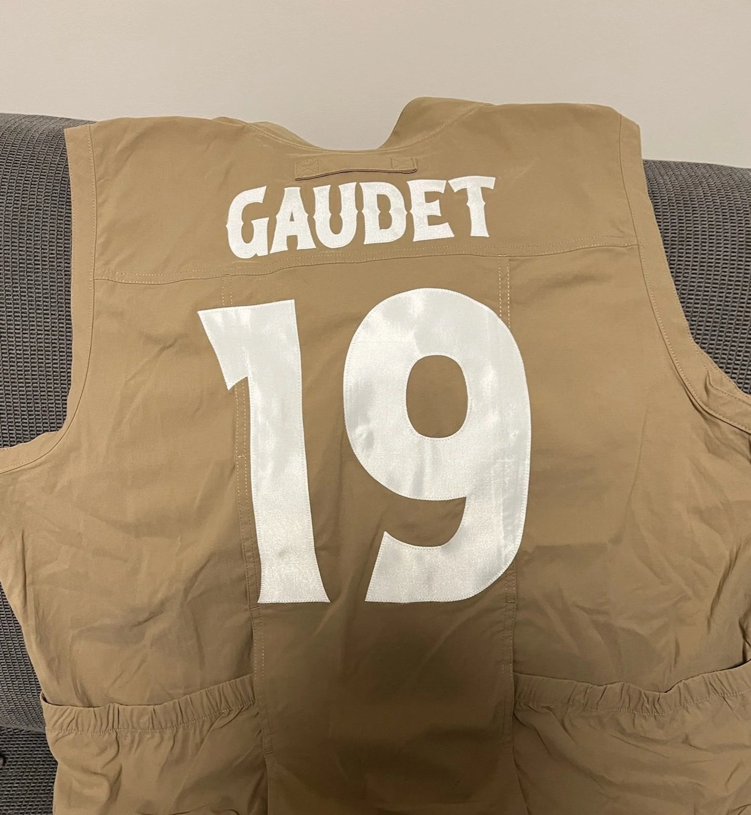 A tan fishing vest with the name 'GAUDET' and the number '19' sewn in white on the back, draped over the back of a dark gray woven fabric couch.