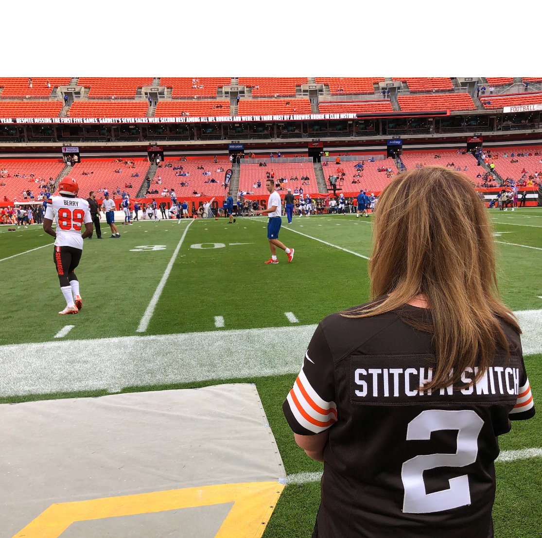 A football stadium with players warming up on the field. A woman with long brown hair, wearing a black Stitch N Switch jersey with the number 2, stands near the sideline watching the game.