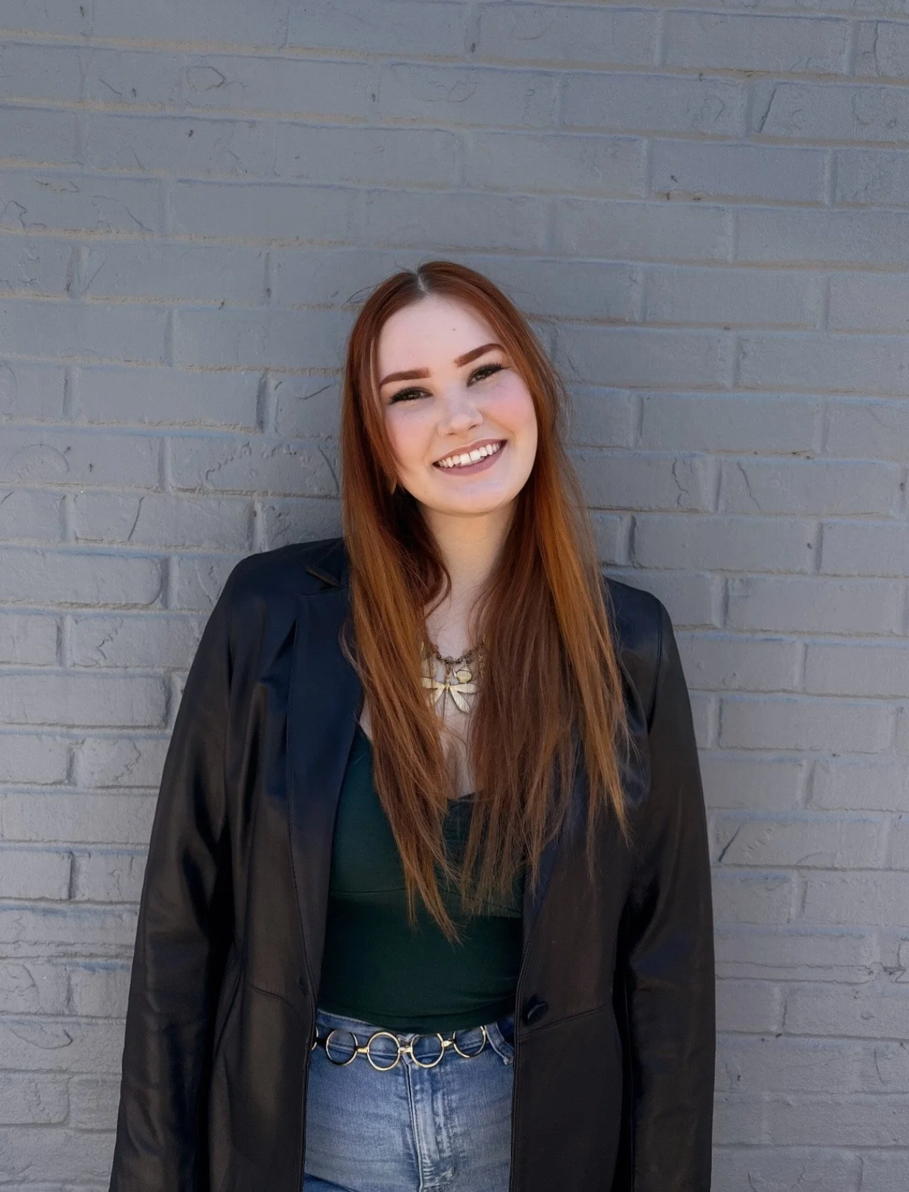 A young woman with long red hair smiling, standing against a gray brick wall. She is wearing a black leather jacket, a dark green top, and blue jeans with a chain belt. She wears makeup with defined eyebrows and lipstick.