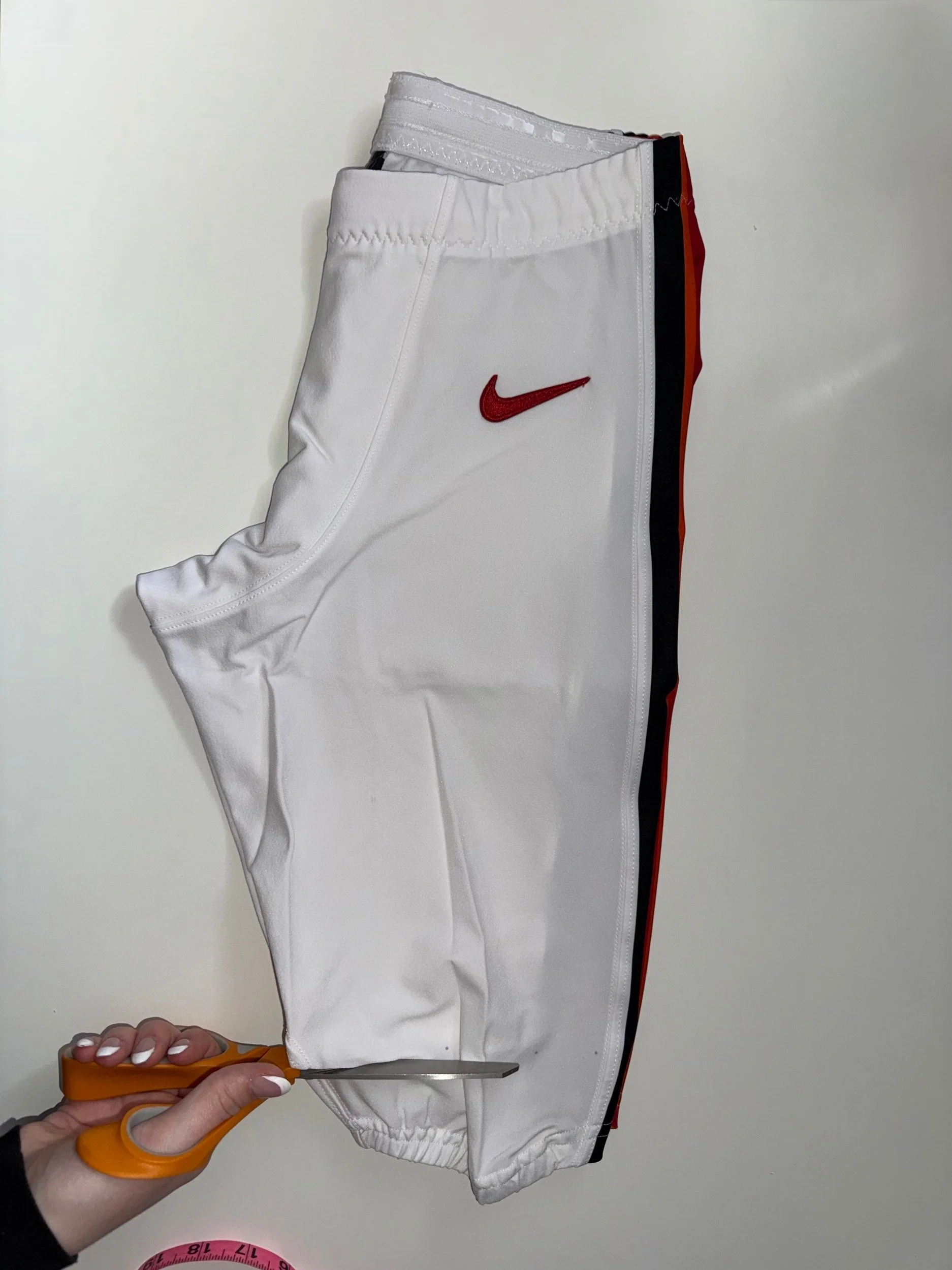 A person is holding a pair of orange scissors and cutting a white football pants with a red Nike logo on it. Tampa Bay Buccaneers.