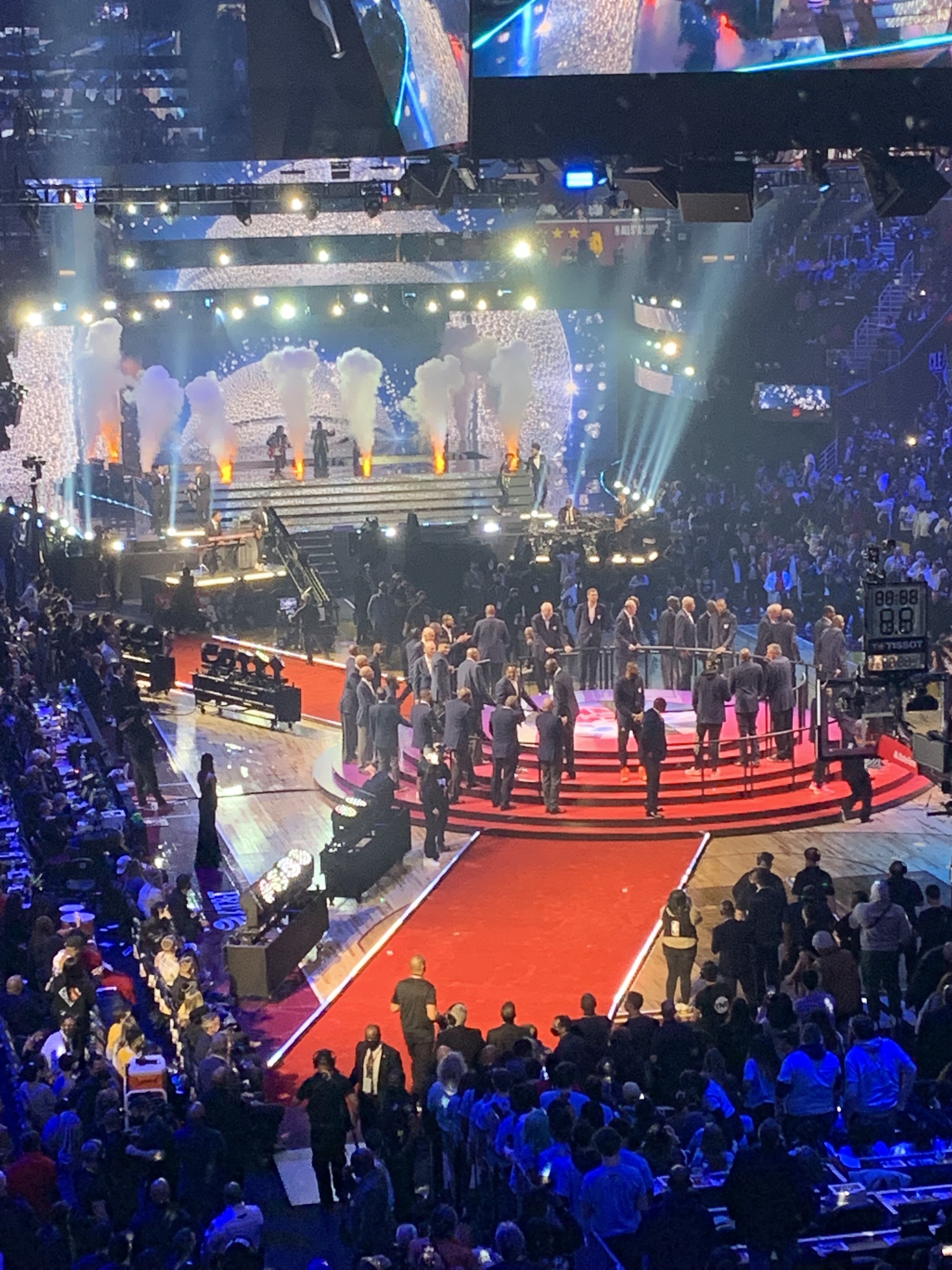 The 75th Anniversary NBA All-Star Game halftime with a red carpet leading to a stage. The stage is illuminated with bright lights, smoke effects, and a large screen displaying visuals. Top players on the red carpet in custom blazers.