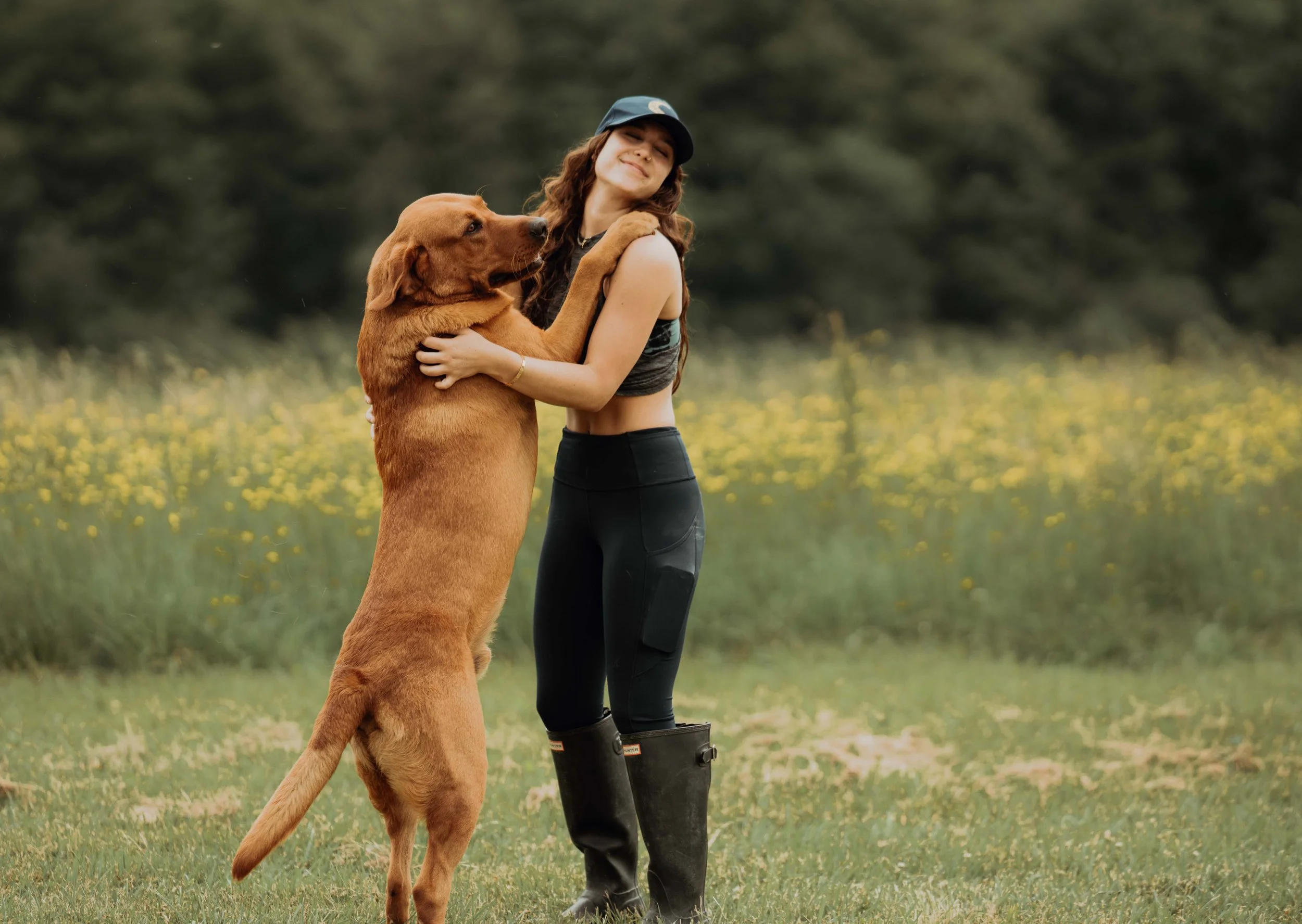 A woman wearing black riding pants and tall black rubber boots hugging a large, brown dog in a grassy field with yellow wildflowers and a forest in the background.