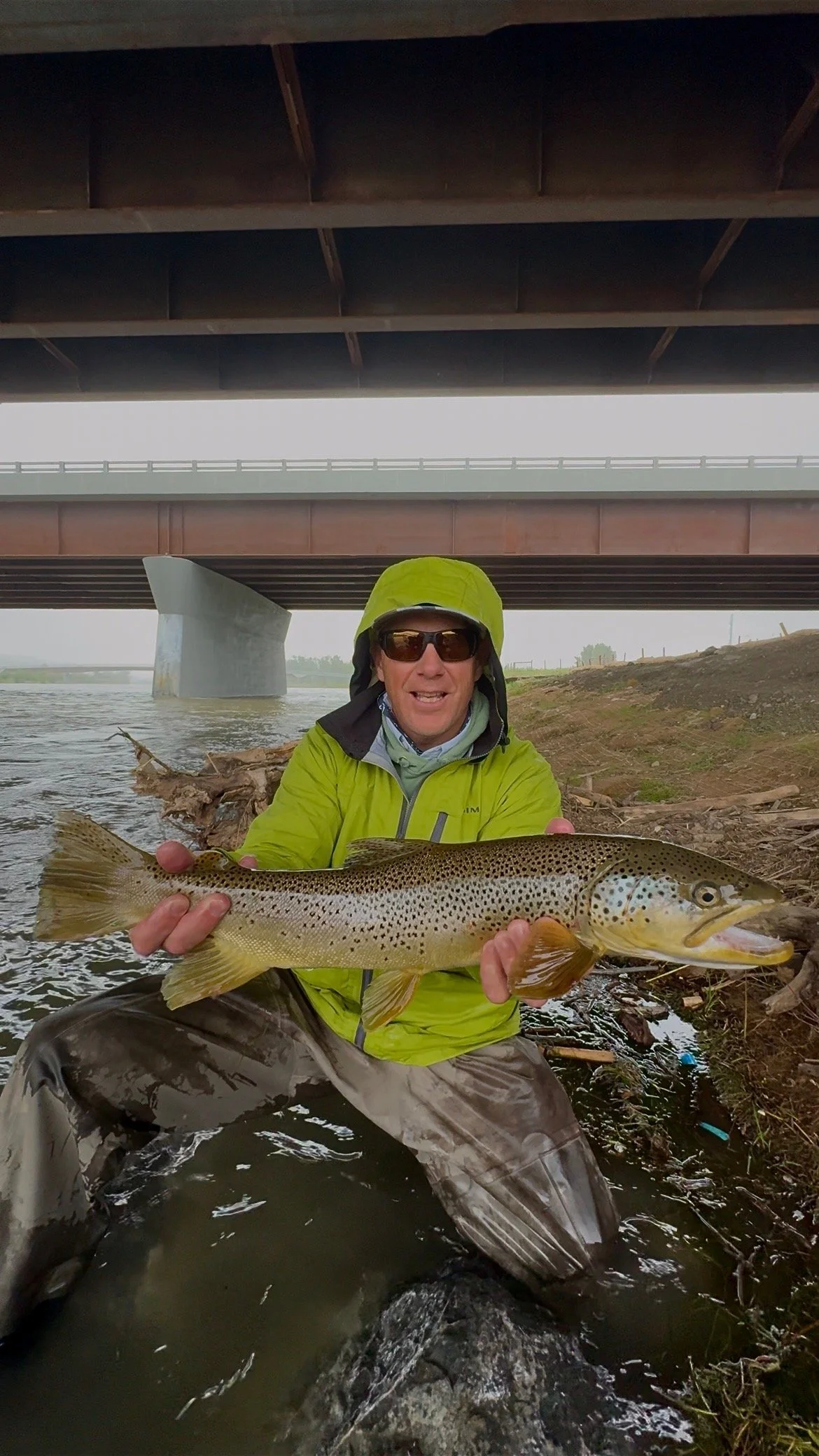 Man wearing sunglasses and a cap, holding a large fish in a river with autumn-colored trees in the background.