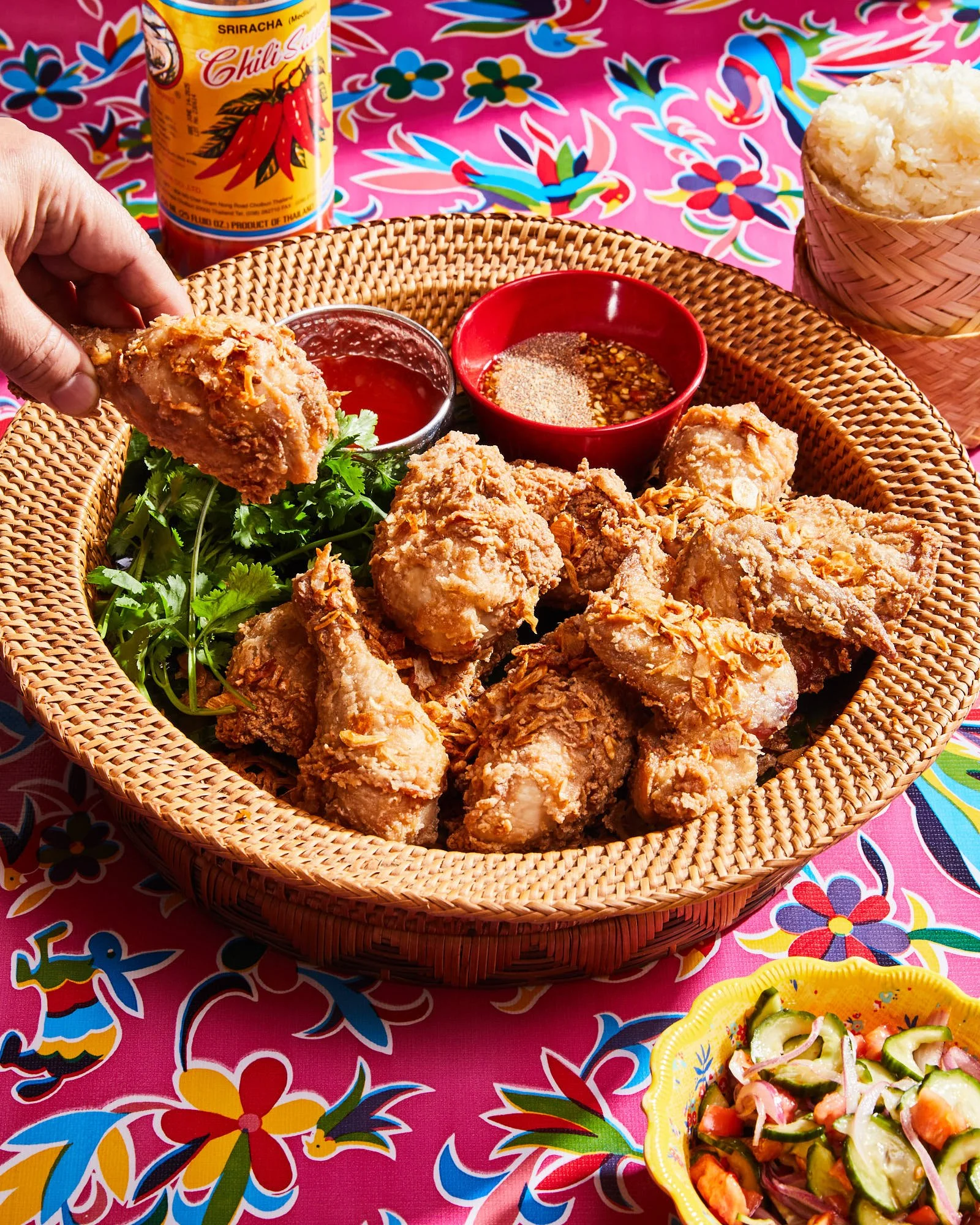 Basket of fried chicken with herbs, dipping sauces, and sticky rice on a vibrant tablecloth; hand holding a piece of chicken.
