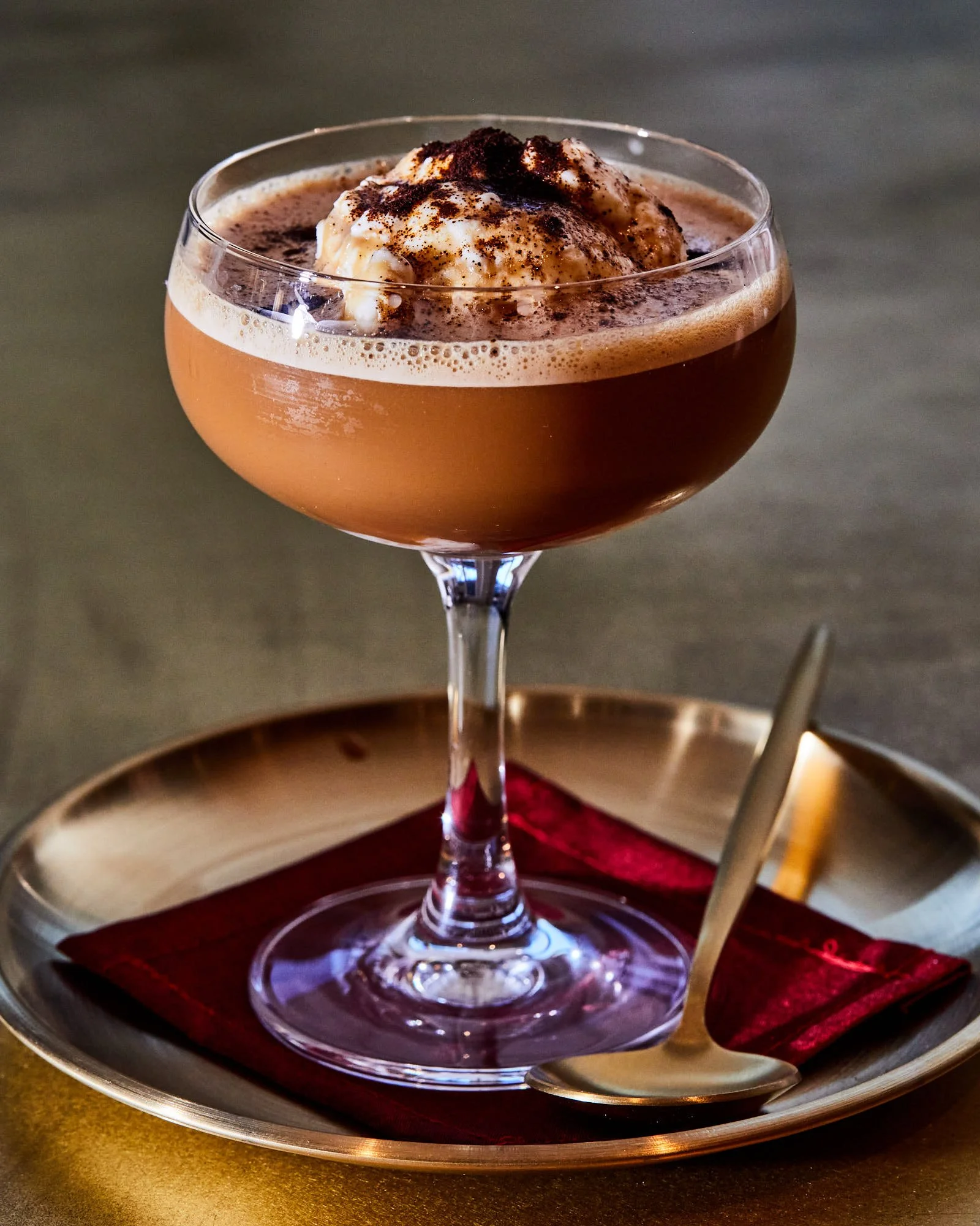 A cocktail glass filled with a affogato topped with whipped cream and chocolate shavings, placed on a golden plate with a red napkin and a spoon.