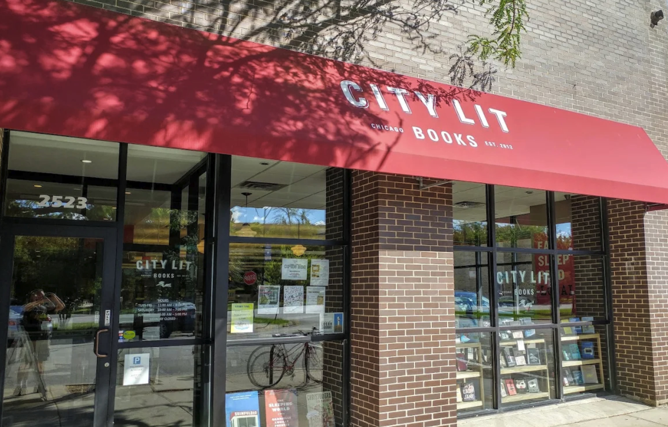 Exterior view of City Lit Books bookstore with a red awning, brick walls, and large glass windows displaying books inside. Reflection of people and bicycles can be seen on the glass.