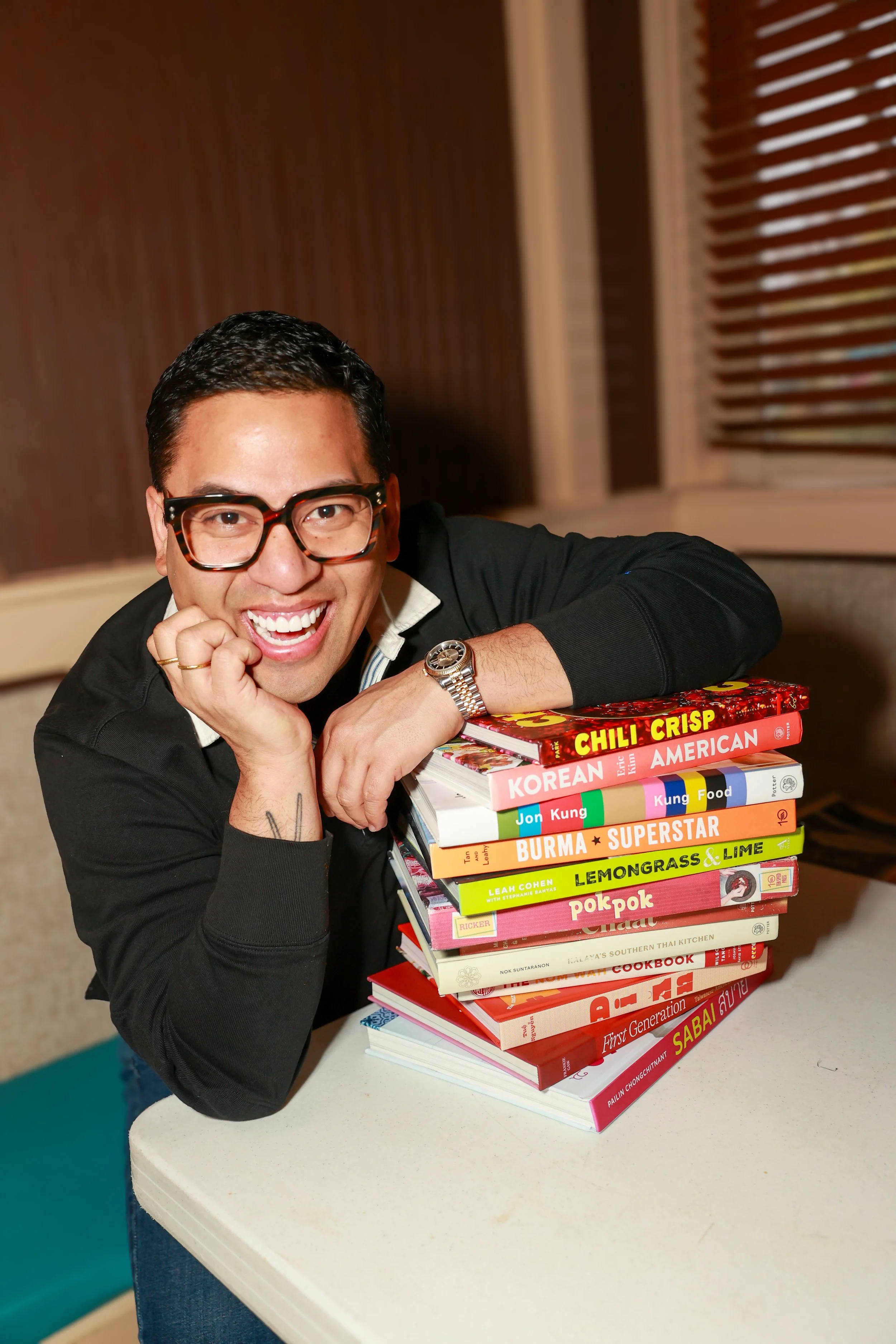 Arnold Myint smiling and leaning on a stack of colorful cookbooks on a table.