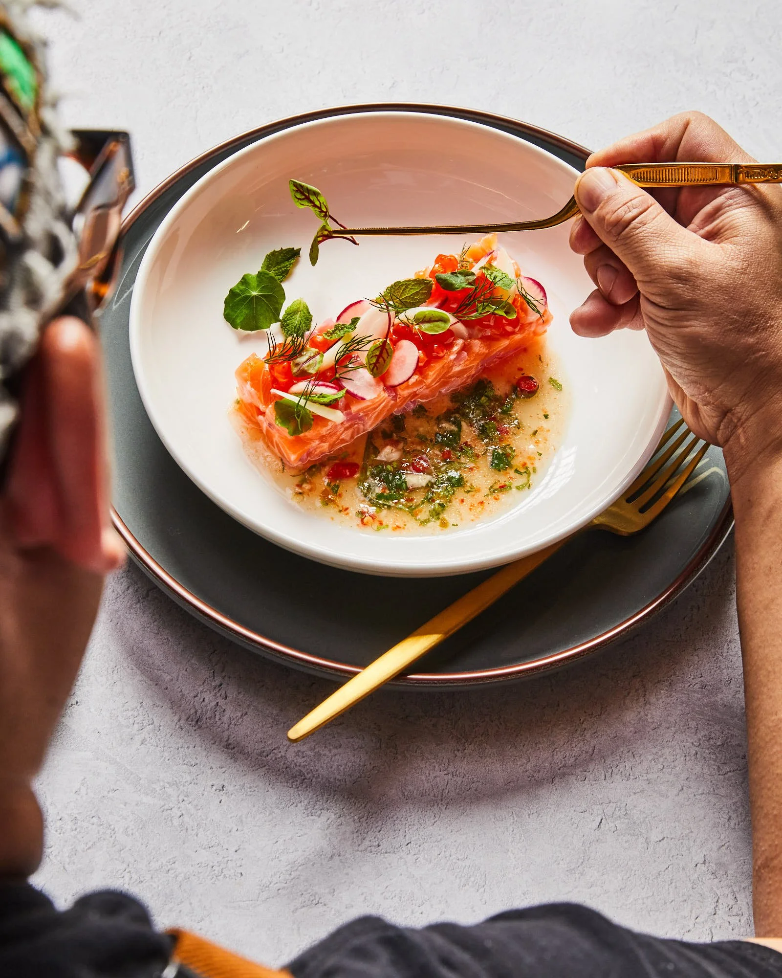 Person holding a fork near a dish with salmon tartare, garnished with herbs and vegetables on a white plate.