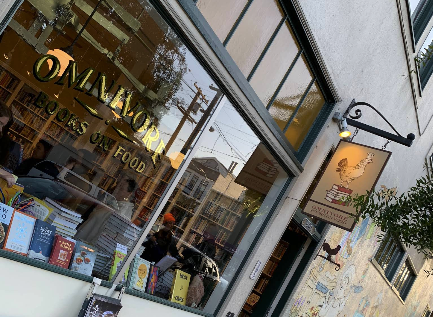 View of a bookstore with large windows, books on display outside, a hanging sign with a chicken sitting on books, inside visible bookshelves, and people shopping.