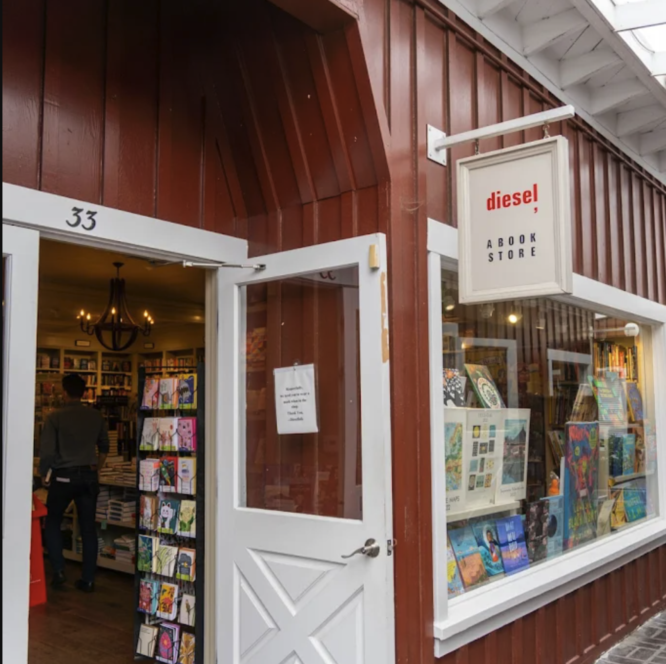 Exterior of a bookstore with a white door slightly open, displaying books inside. A hanging sign reads 'diesel, A BOOK STORE' in red and black letters. The store has a red exterior with large windows showcasing colorful books inside.