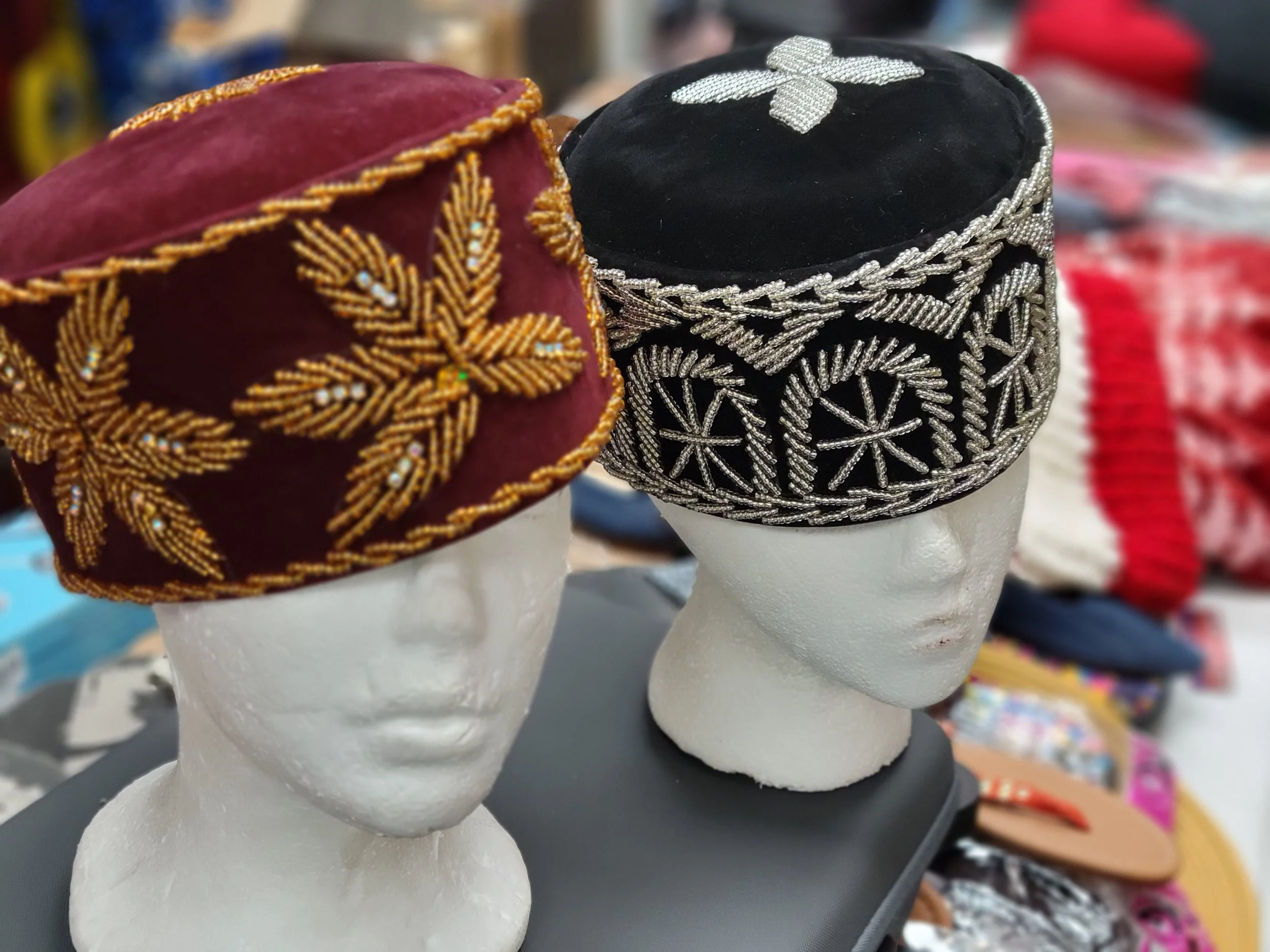 Two mannequin heads wearing ornate traditional caps, one burgundy with gold embroidery and the other black with silver embroidery, displayed on a table with various textiles in the background.