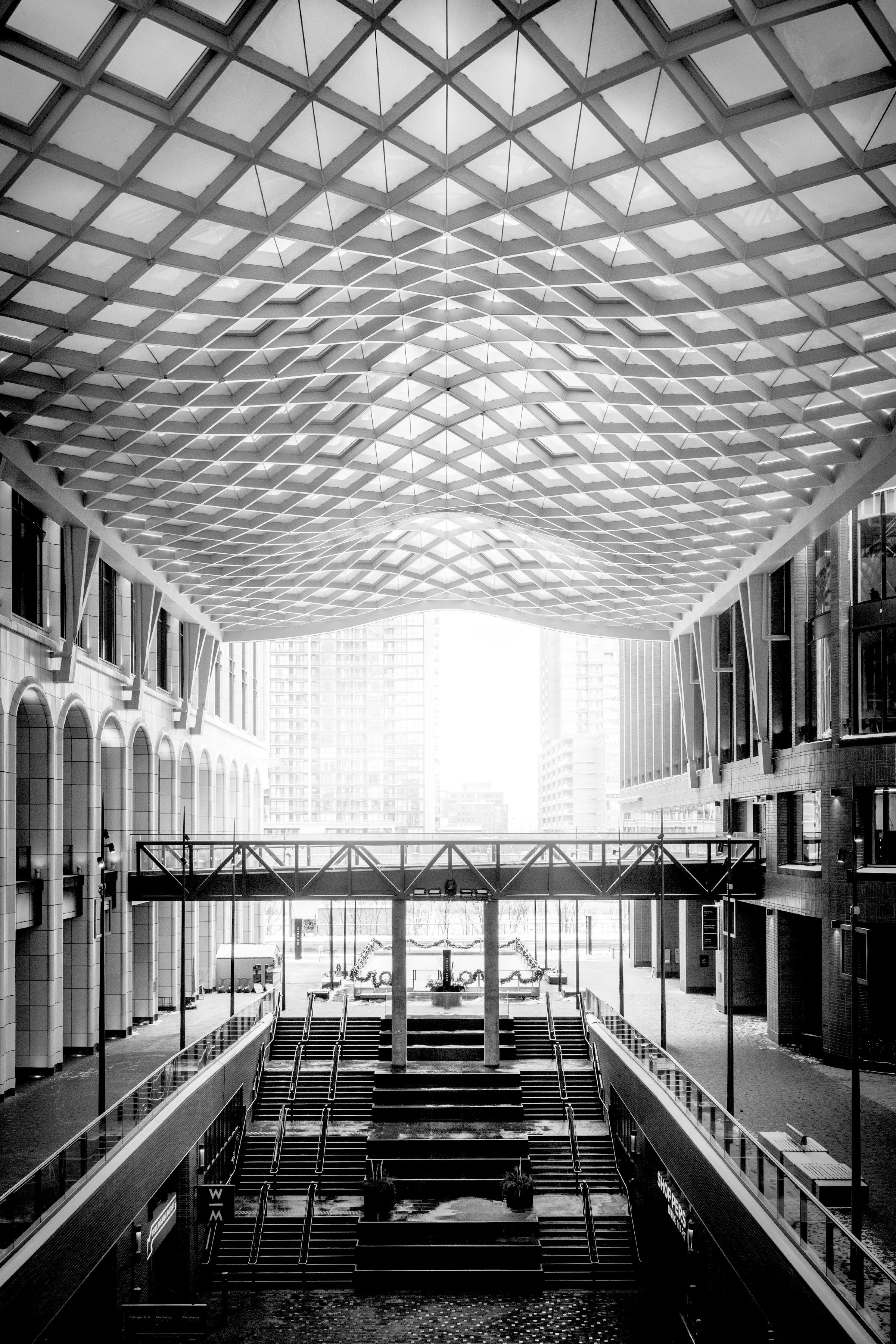Modern indoor urban space with a high patterned ceiling, stairs, and city buildings visible through large windows.