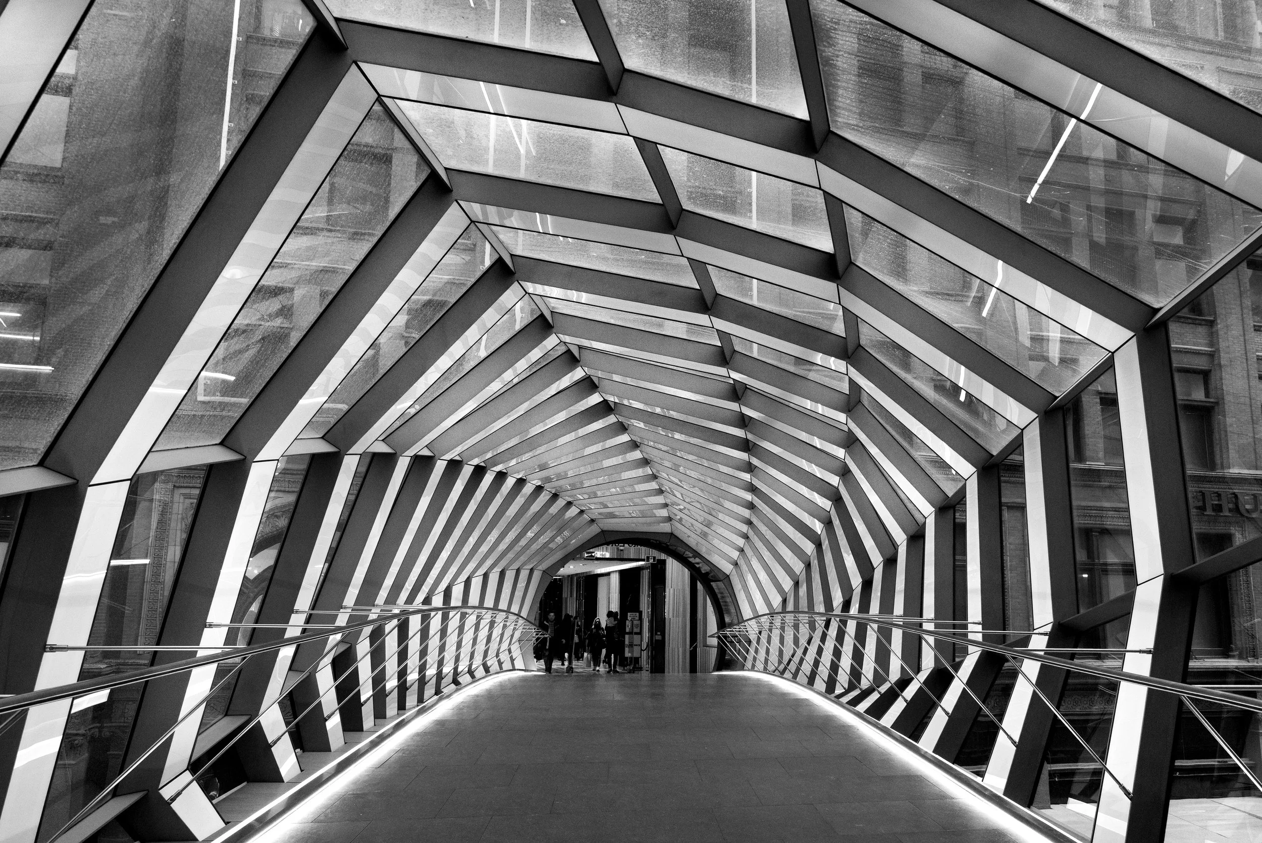 Interior view of a modern architectural pedestrian bridge with a geometric, tunnel-like design of metal and glass, leading to a city building entrance.