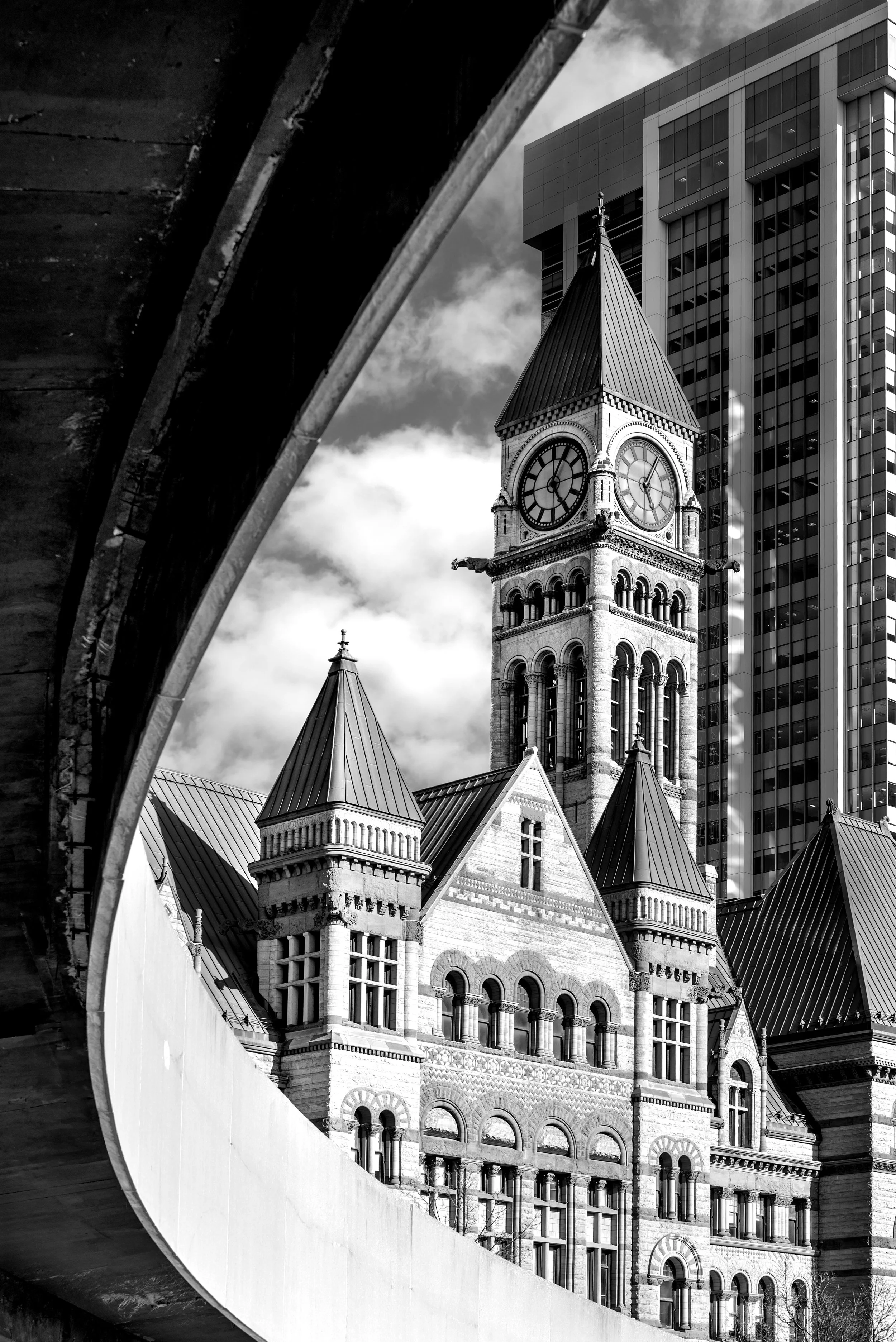 Black and white photo of a historic clock tower with Roman numerals, surrounded by modern high-rise buildings, seen through an arched bridge or structure.