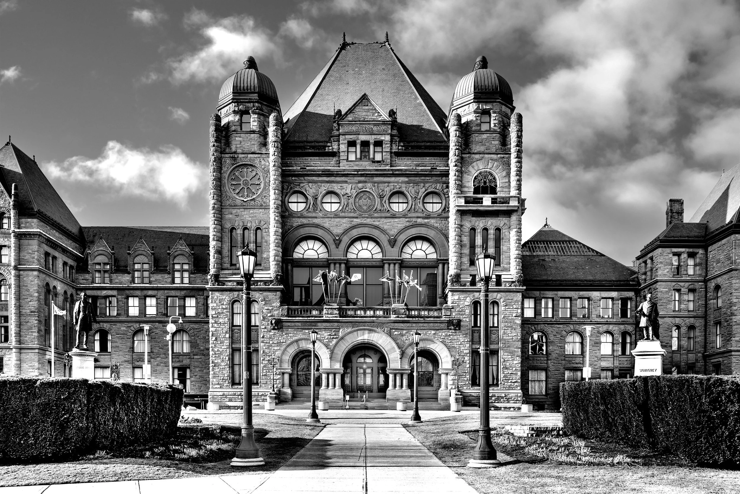 Black and white photo of a large historic stone building with turrets and a steep roof, featuring flags, statues, and street lamps in front.