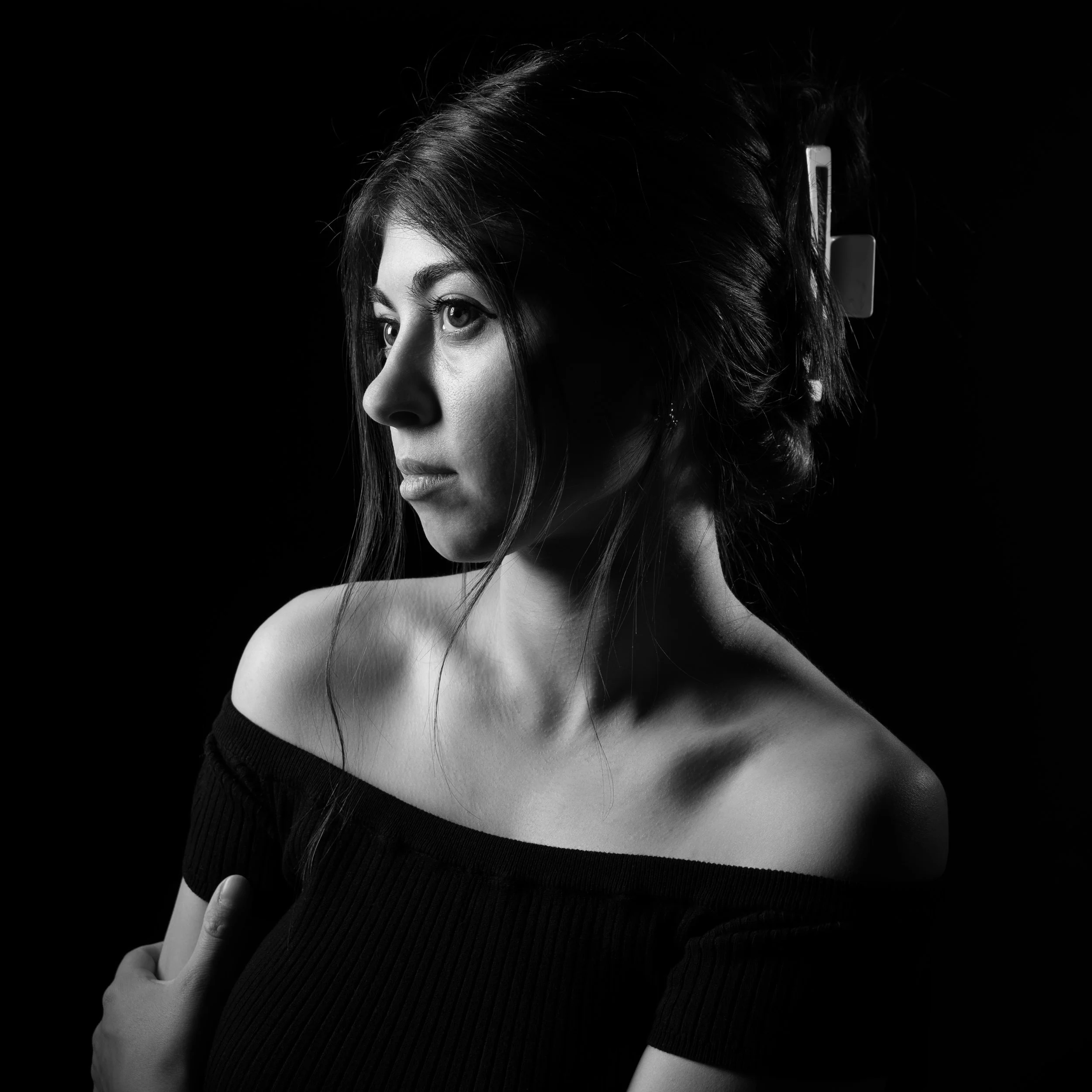 Black and white portrait of a young woman with long dark hair, wearing an off-the-shoulder top, looking to the left, with soft lighting highlighting her face and shoulder.