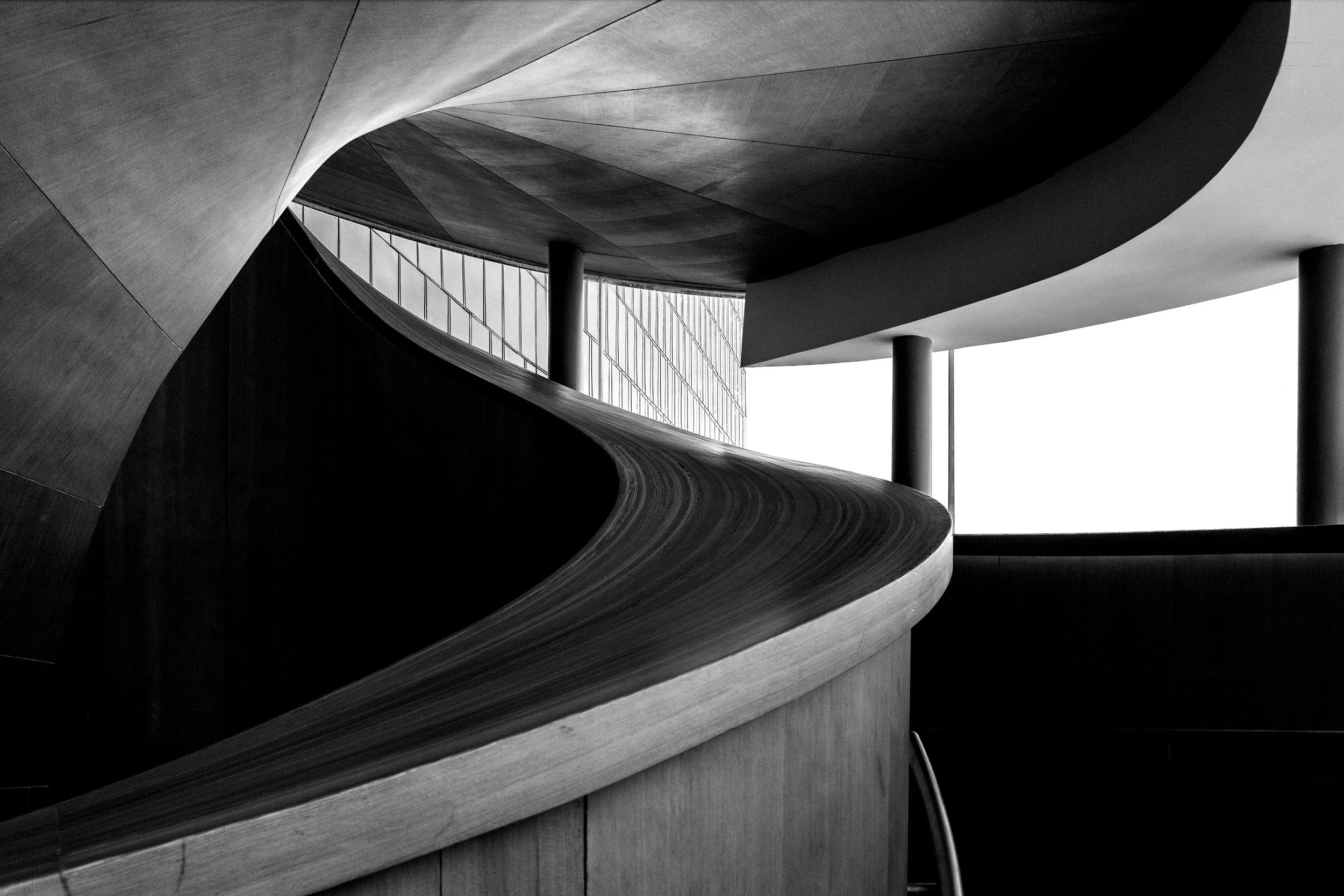 Black and white photo of modern architectural interior with curved wooden staircase, large windows, and supporting columns.