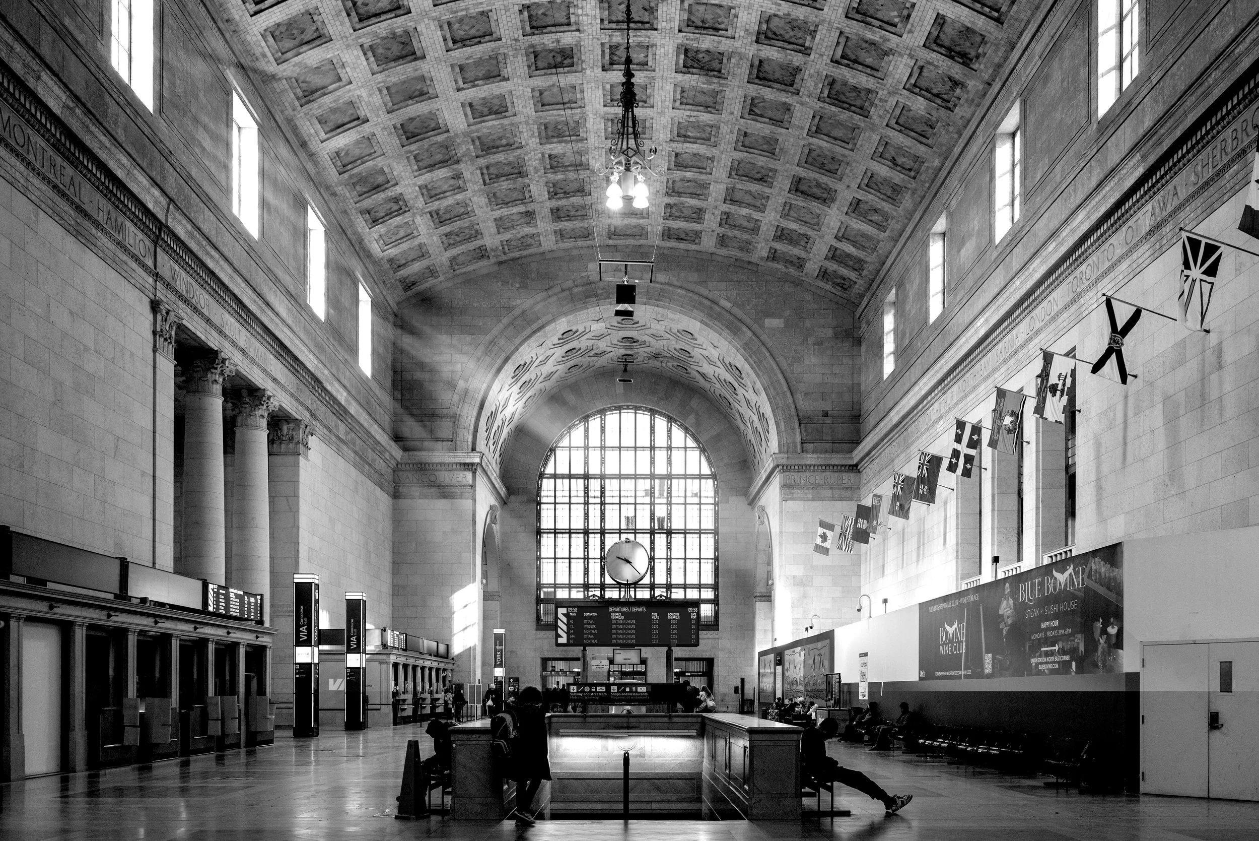 Interior view of a historic train station with high arched ceiling, large clock, and ticket counters, with people sitting and waiting.