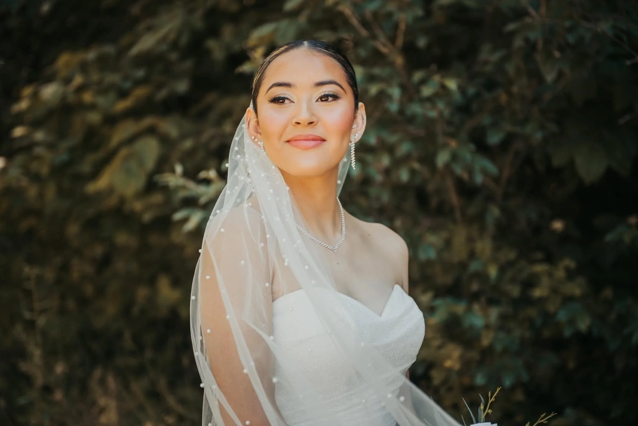 A bride wearing a white strapless wedding dress and a delicate veil with pearls stands outdoors in front of greenery, smiling softly.