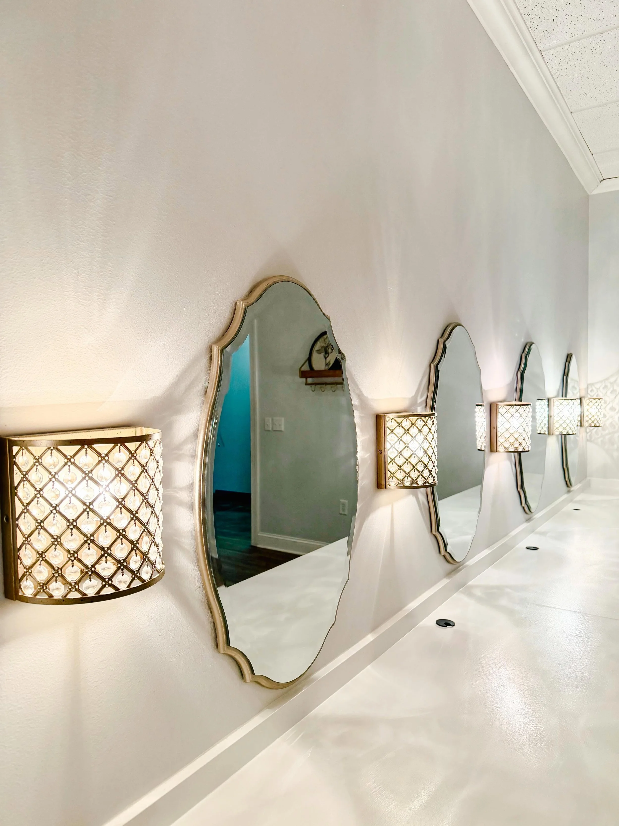 Multiple decorative mirrors mounted on a white wall, each with unique frames, and stylish wall sconces with patterned lampshades between the mirrors.