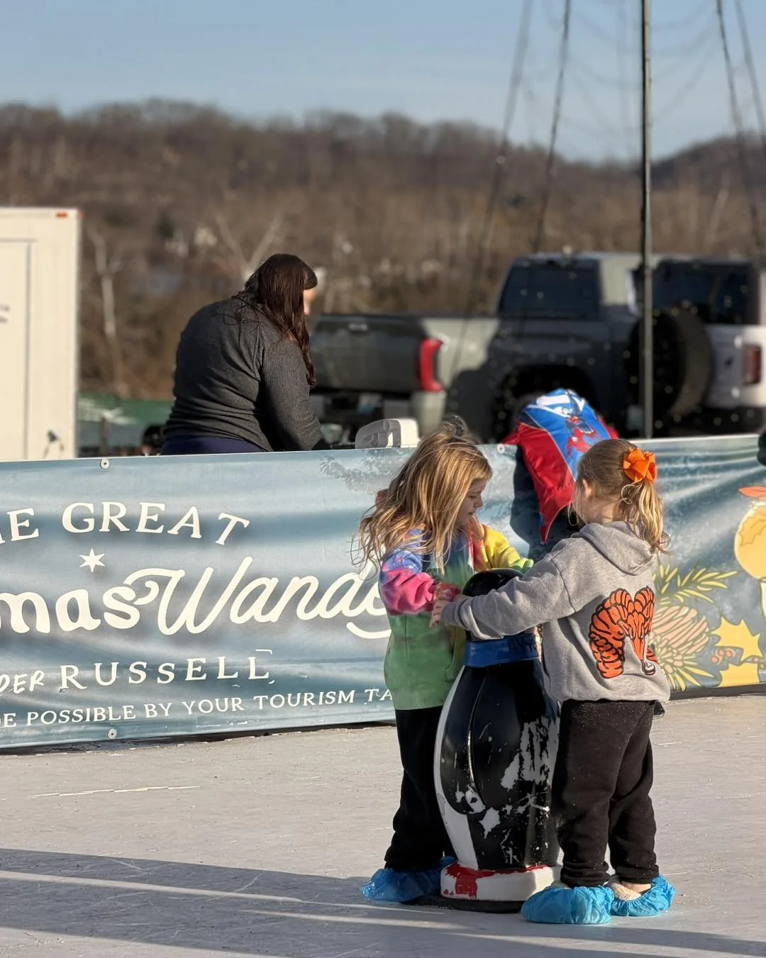 Two children wearing blue shoe covers are holding a cylinder shape object, with a woman in a black jacket and two trucks in the background. A banner with the words 'The Great Christmas Wonderland' is also visible.