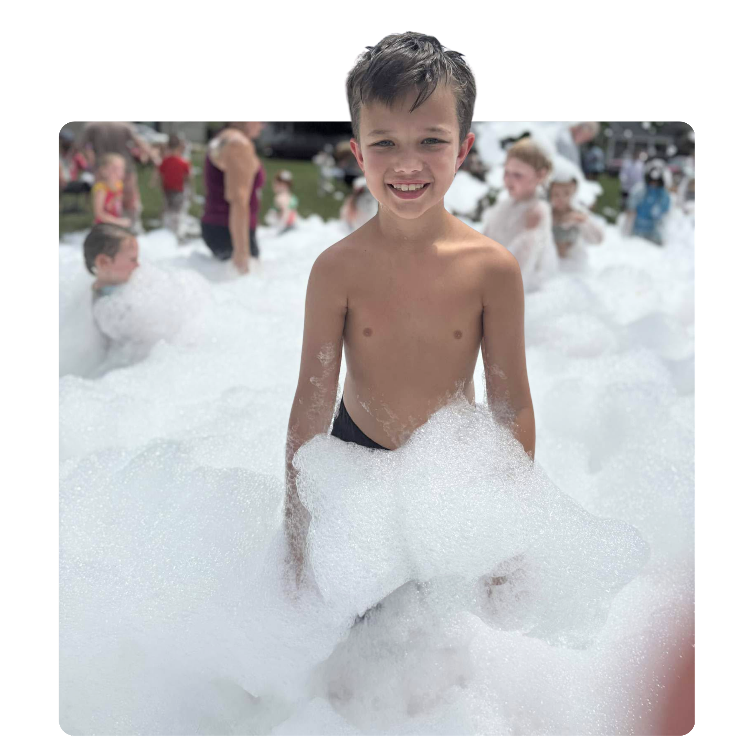 A young boy with wet hair smiling in the foreground, standing in a foam-filled outdoor area with other children and adults in the background.