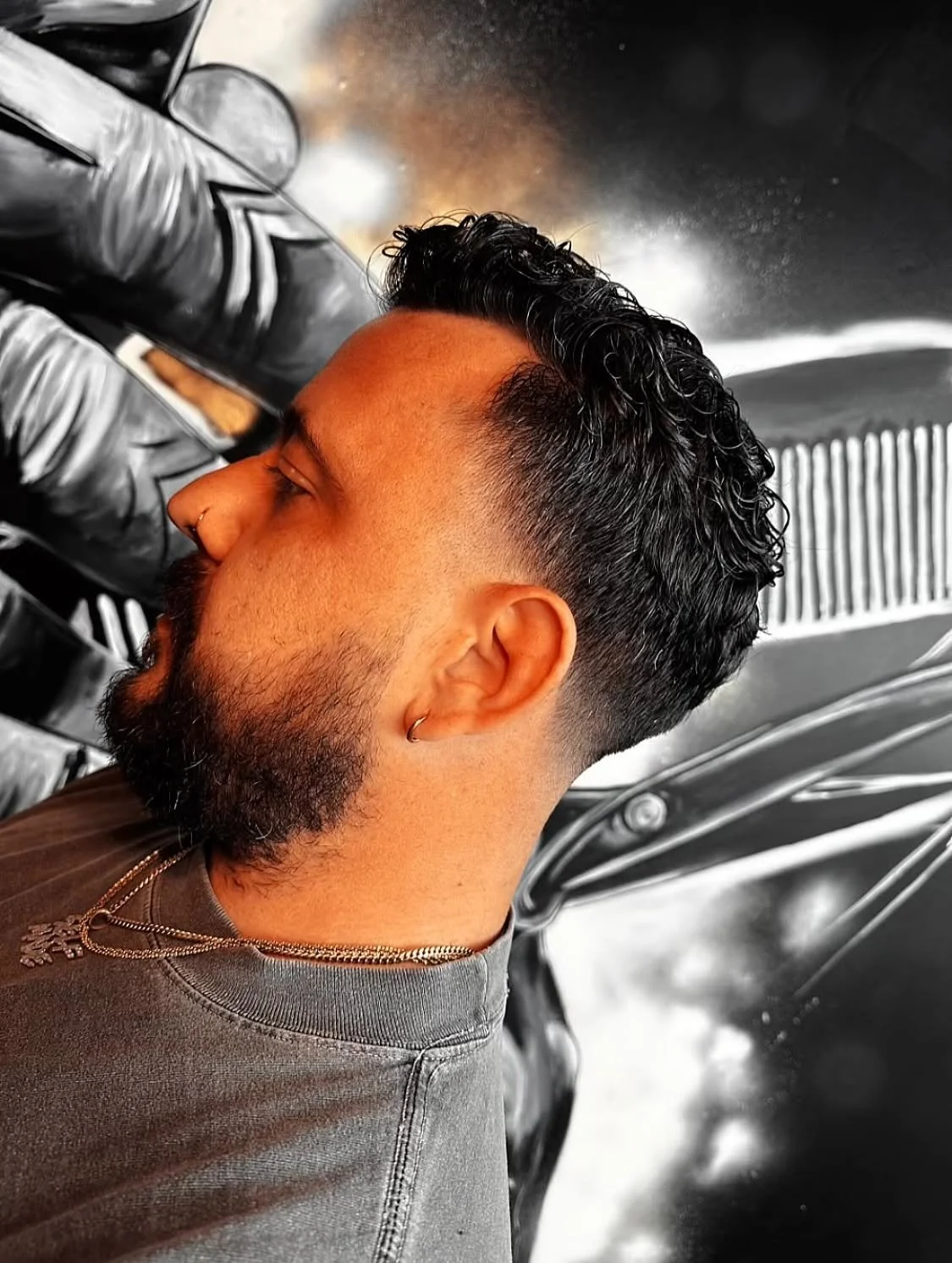 A man with dark, curly hair and a beard receiving a haircut at a barbershop, with black and white decor in the background.