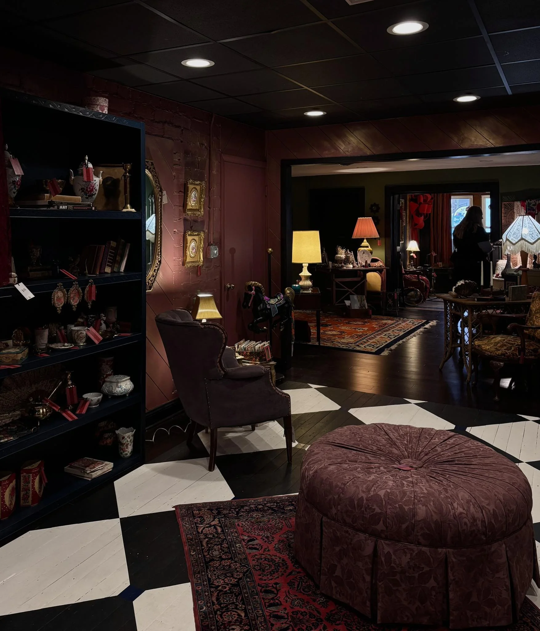 A cozy vintage-style living room with dark wood flooring, red and black patterned carpet, plush purple armchair, and large ottoman. Shelves and lamps illuminate the space, and a person stands near a window in the background.