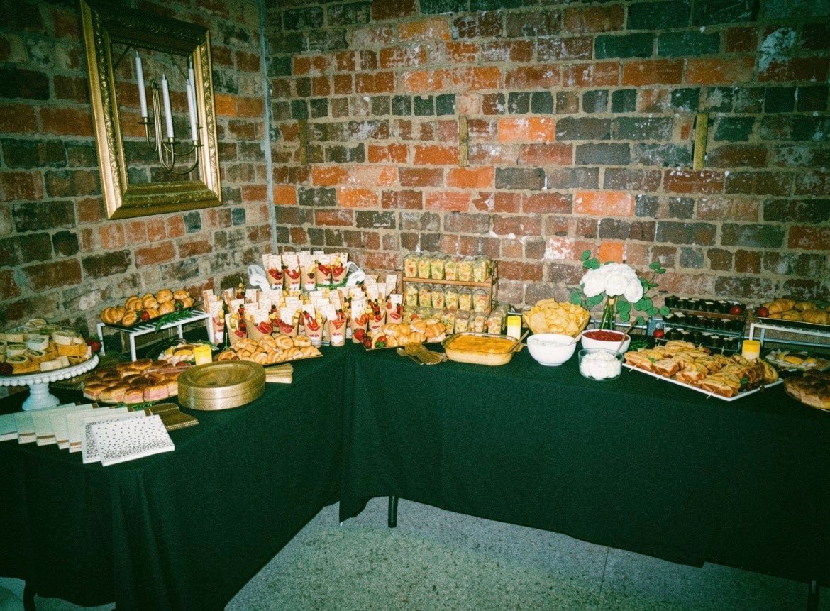 A buffet table with various finger foods including sandwiches, appetizers, and desserts, set against a brick wall with a framed mirror and decorative candelabra.
