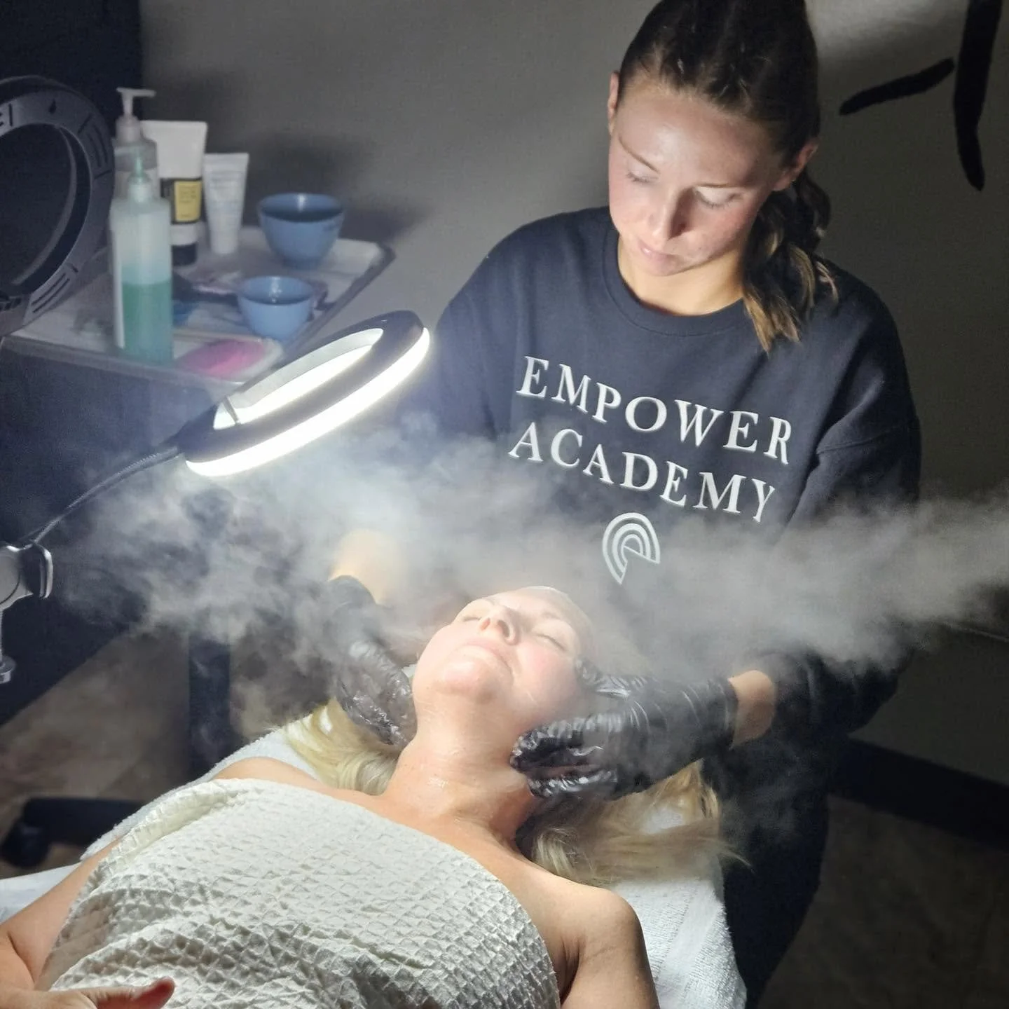 A woman with blonde hair is receiving a facial treatment in a spa or skincare clinic, lying on a treatment bed with her eyes closed. A skincare professional wearing a black sweatshirt with the words 'EMPOWER ACADEMY' is standing beside her, applying a treatment. There is a ring light illuminating the woman's face, surrounded by mist or vapor.