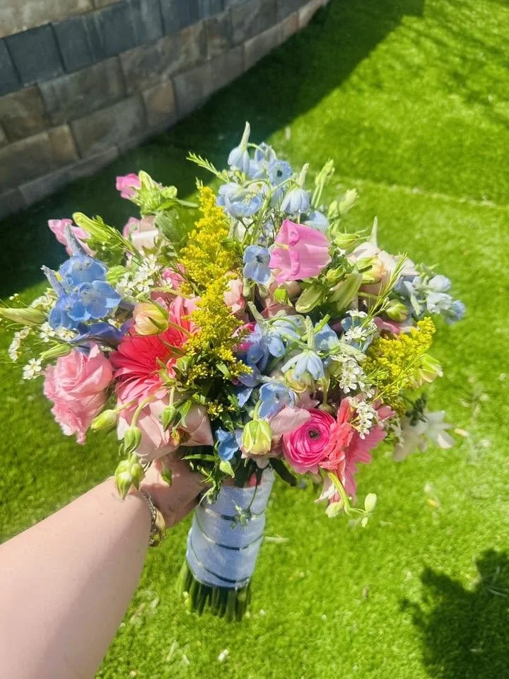 A person holding a vibrant bouquet of mixed flowers outdoors with a green lawn and brick wall background.
