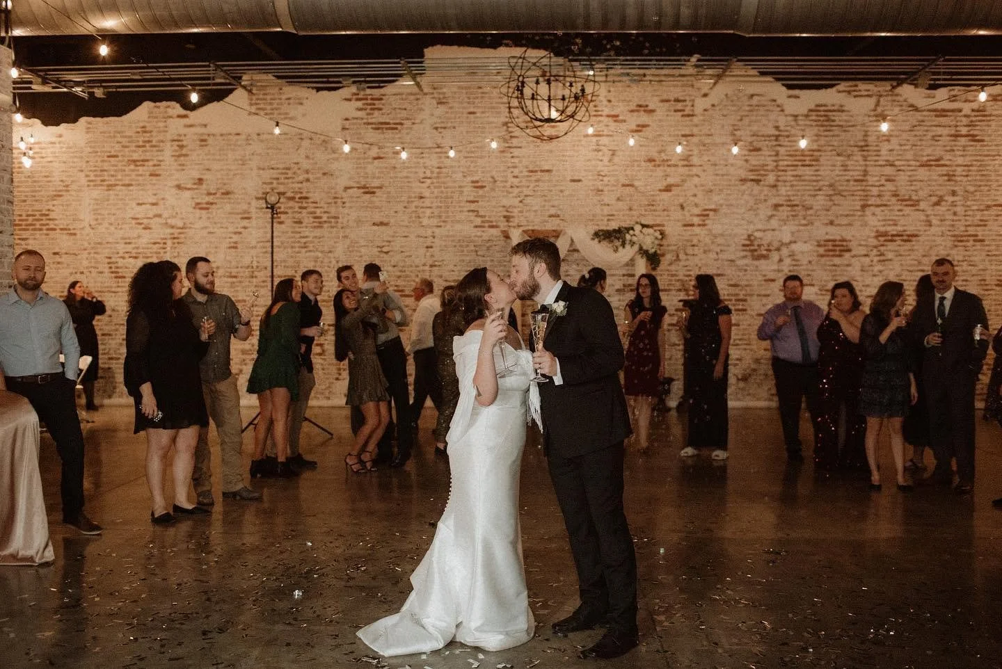 A bride and groom share a kiss on the dance floor at their wedding reception, surrounded by guests dancing and celebrating in a rustic venue with exposed brick walls and string lights.
