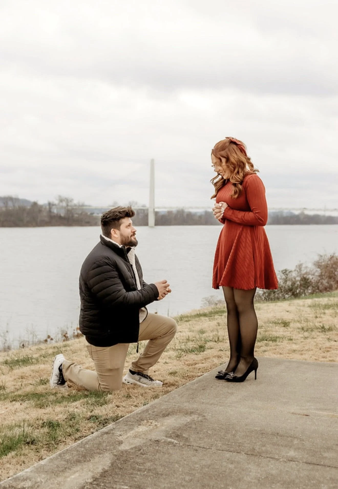 A man kneeling down on one knee proposing to a woman standing in front of him by a body of water with a bridge in the background, on a cloudy day.